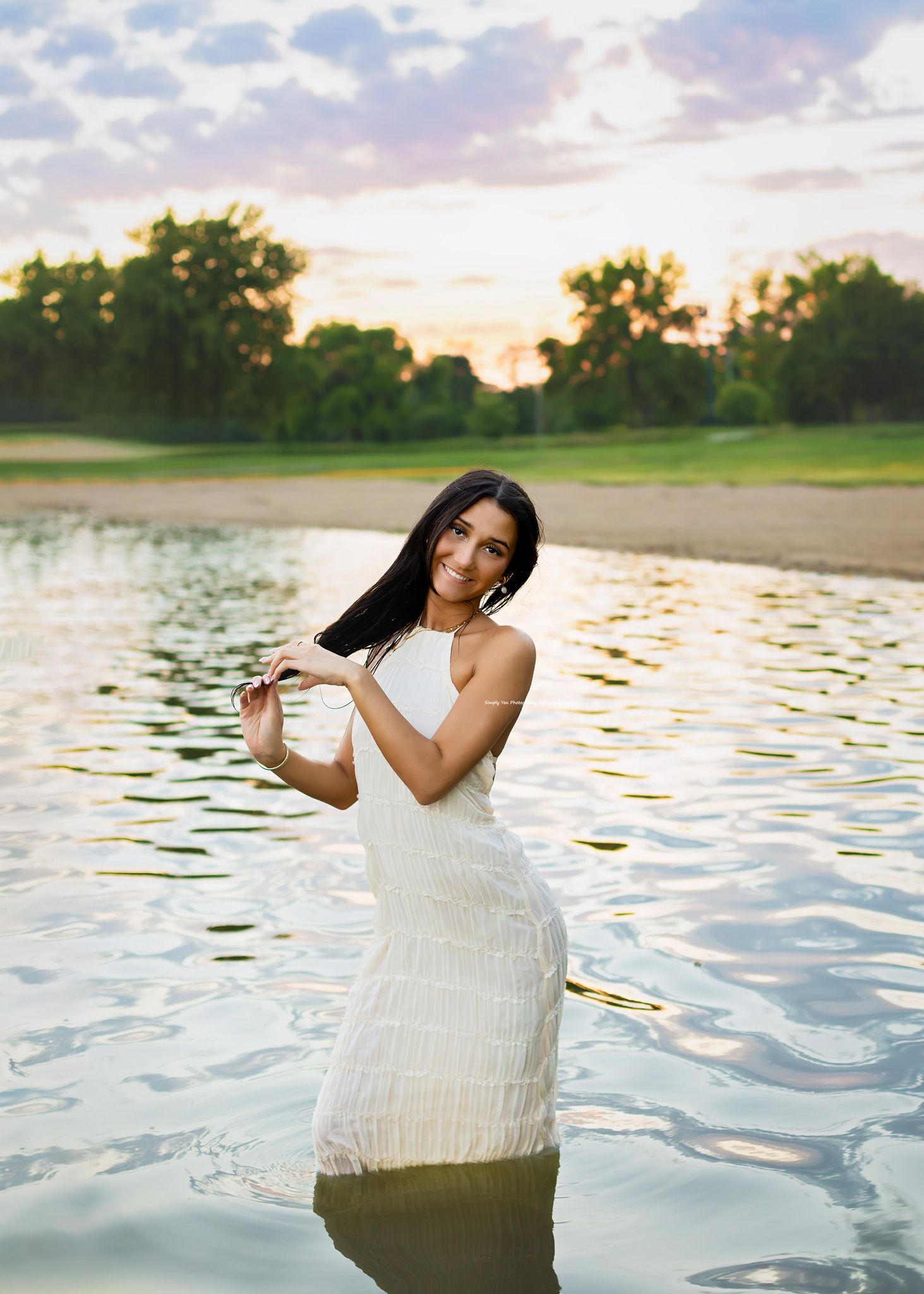 Smiling young woman in white fitted halter dress standing in lake at colorful sunset, senior portrait.
