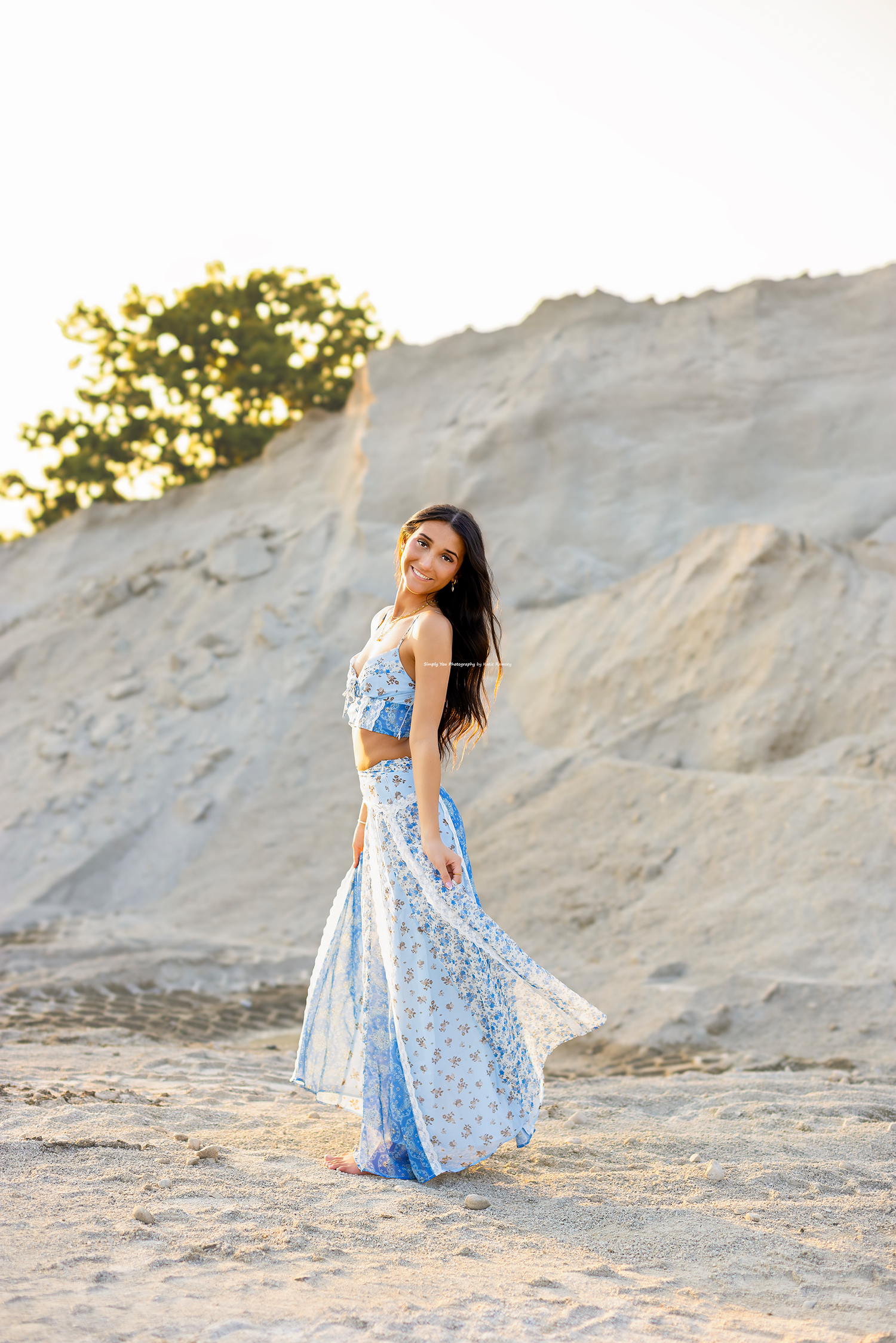 Young woman in blue floral two-piece outfit poses gracefully on sandy dunes at golden hour.