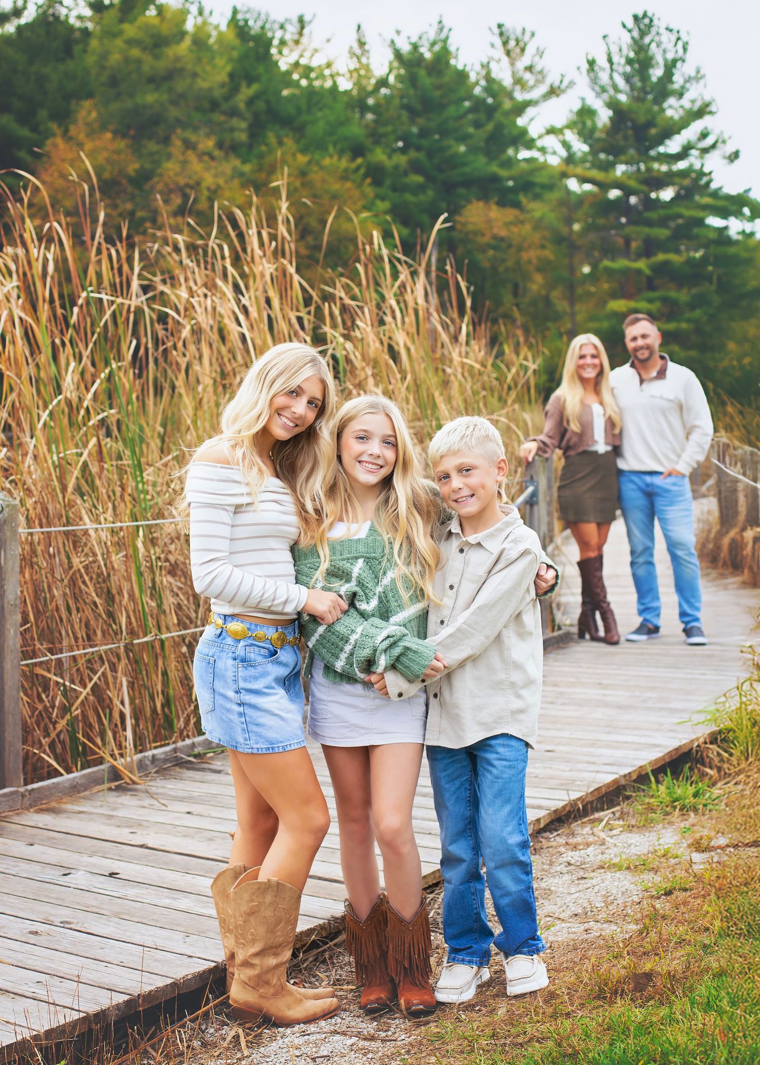 Three siblings pose on a boardwalk while parents stand behind them among tall autumn grasses.