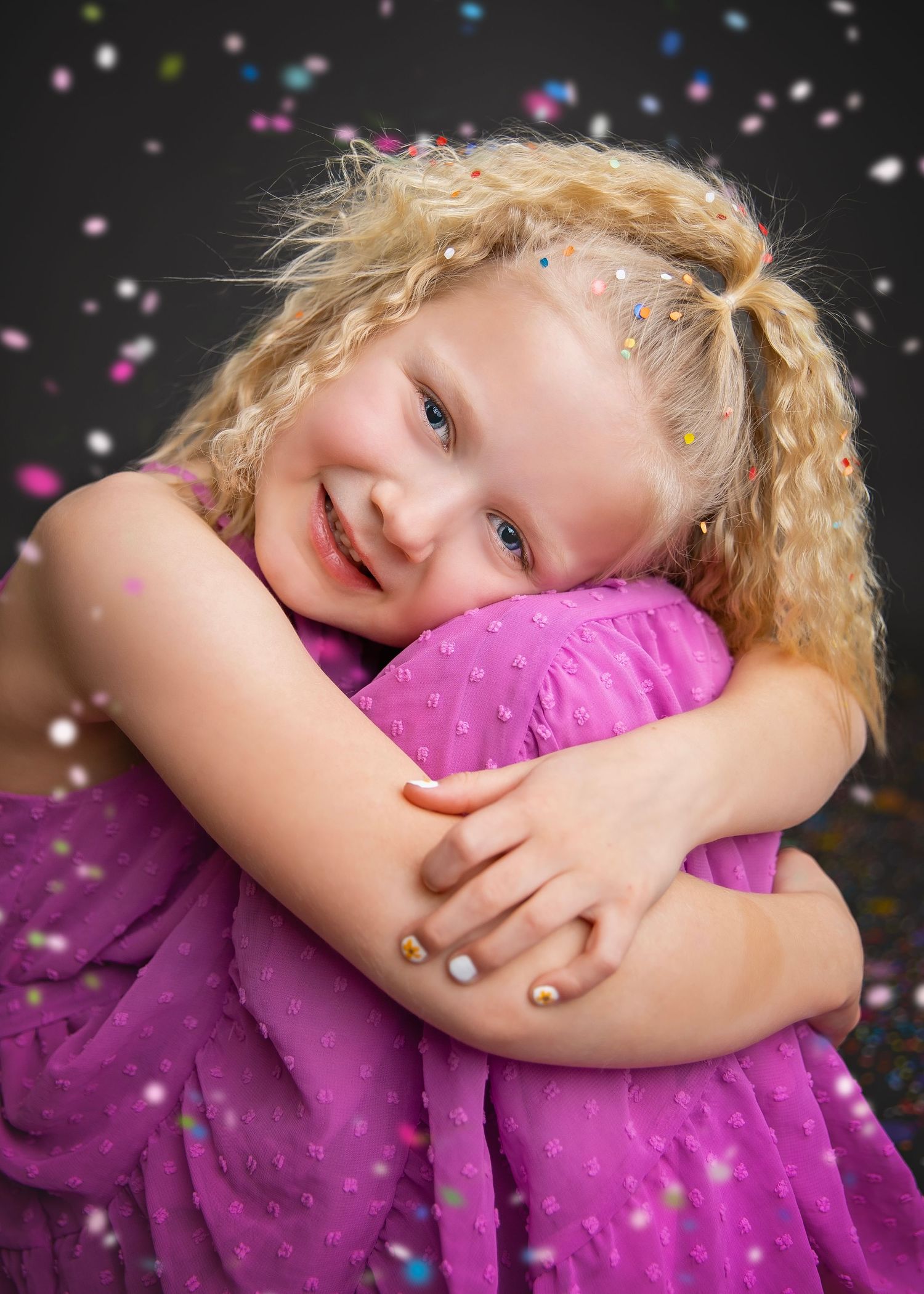 Happy blonde girl in pink dress hugging herself with colorful confetti on dark background.
