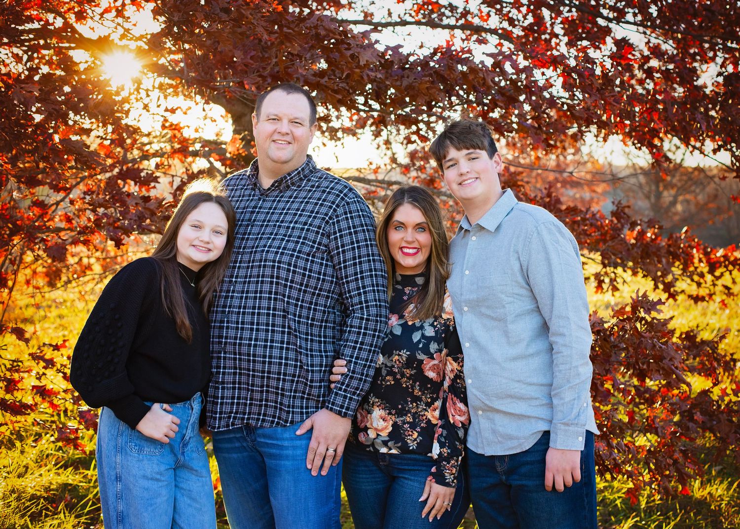 A family of four smiles together under brilliant red autumn leaves during a vibrant outdoor fall photo session.