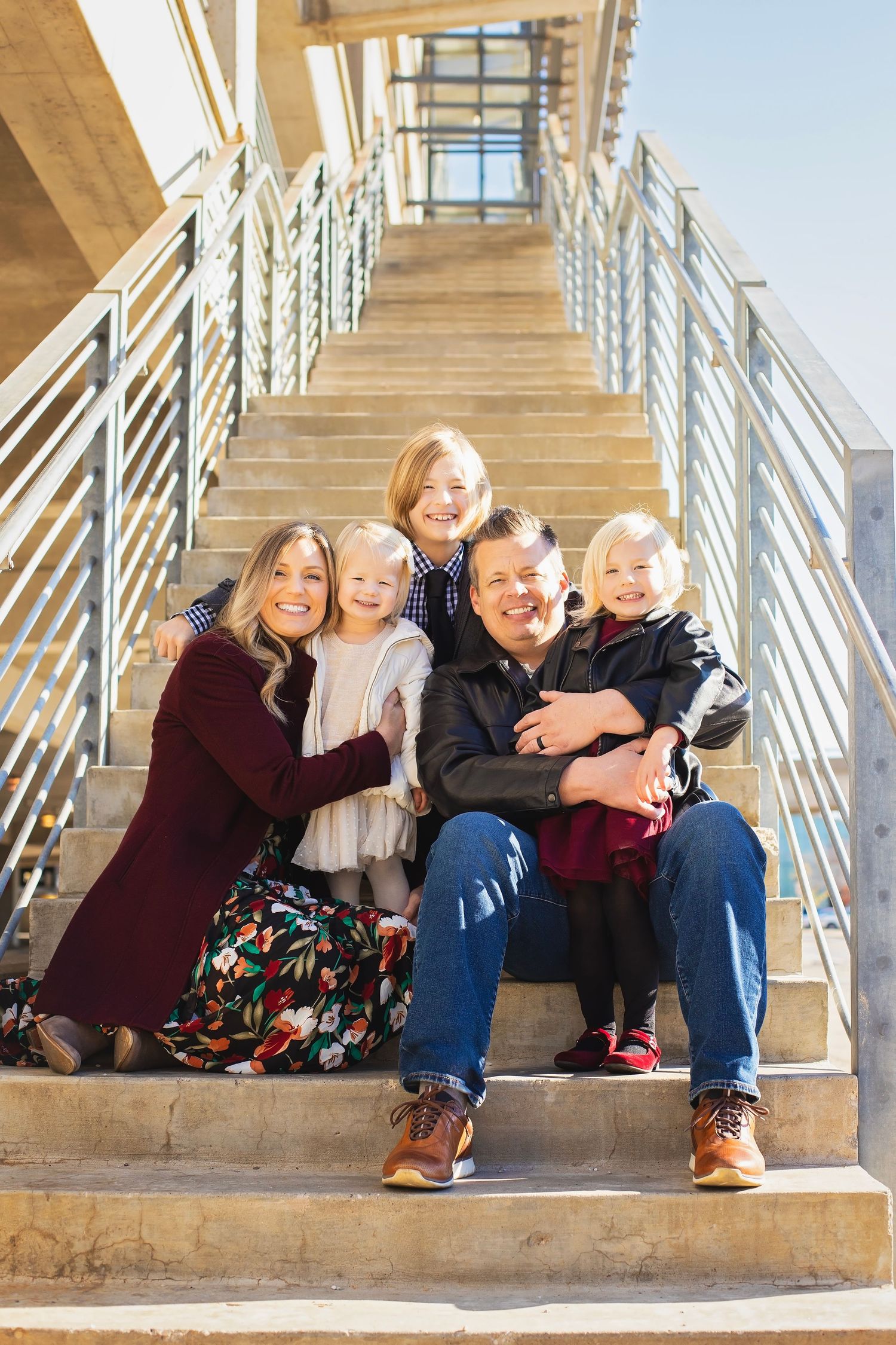 A joyful family of five sits closely together on outdoor stairs, smiling in bright natural sunlight.
