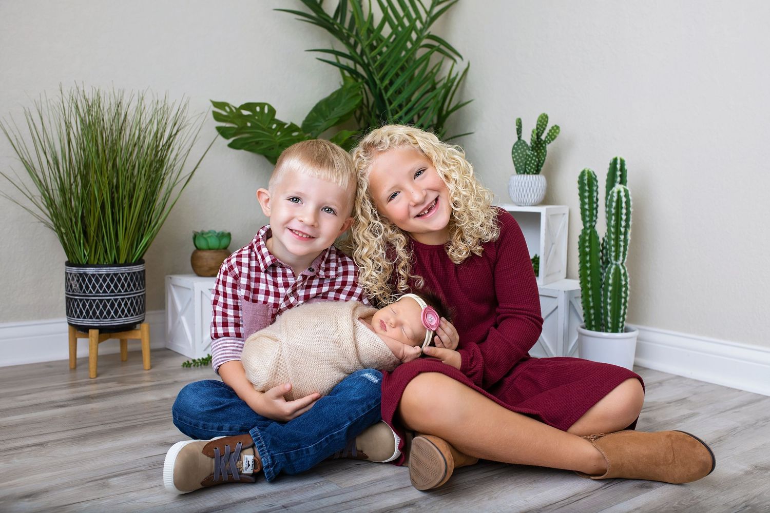 Young boy and curly-haired girl sitting on floor holding sleeping newborn baby with cacti backdrop.