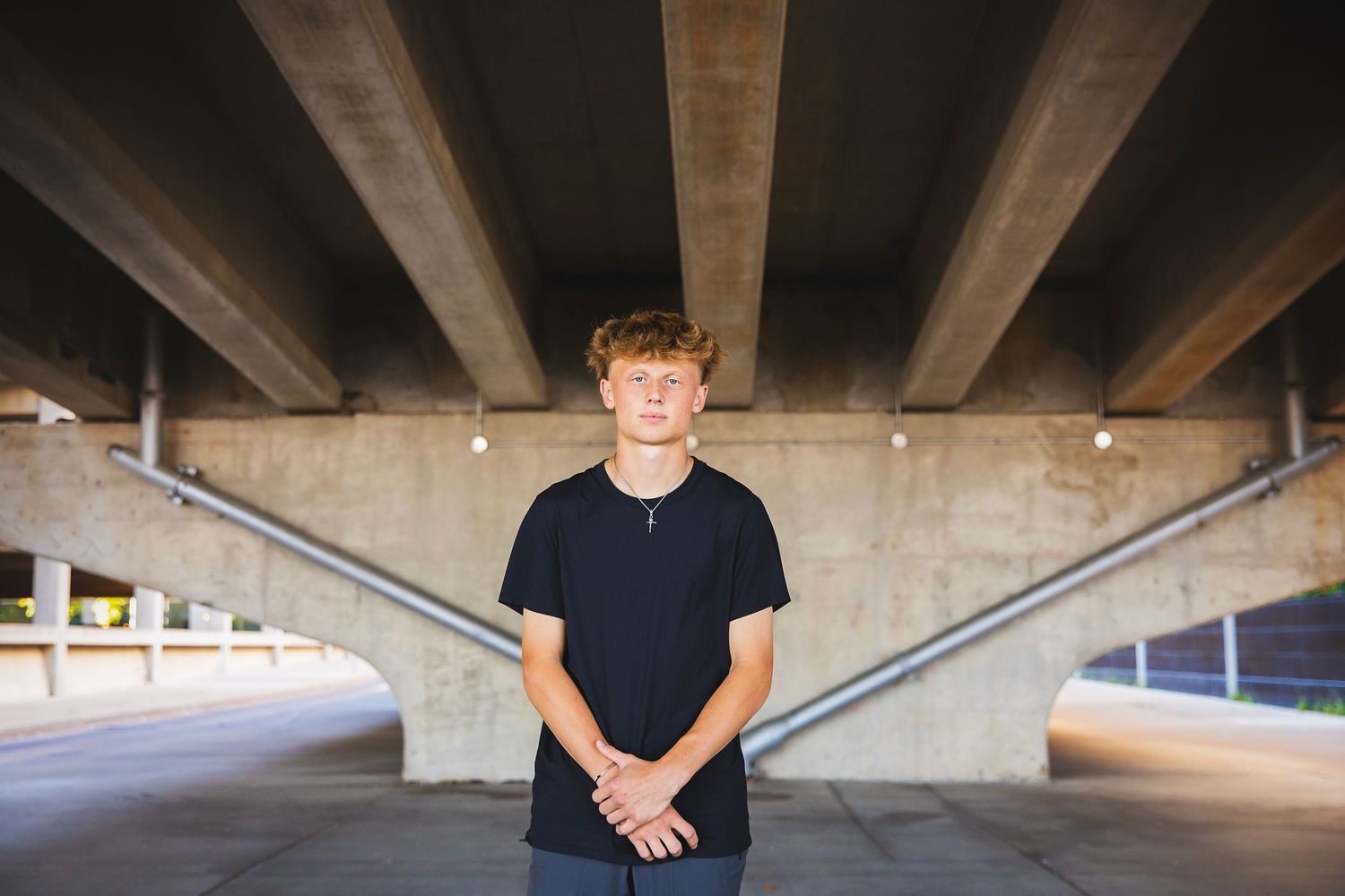 Teen boy in black t-shirt standing under concrete bridge overpass with hands clasped in front.