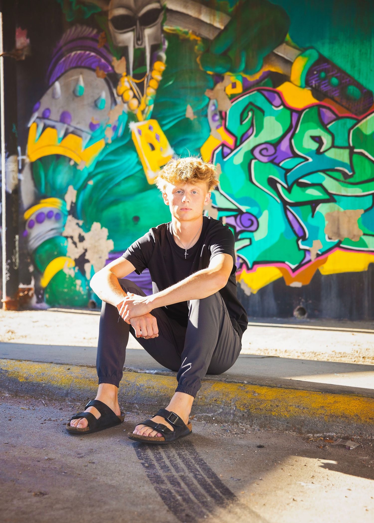Teen boy with curly hair sits relaxed on ground in front of colorful green graffiti mural wall.