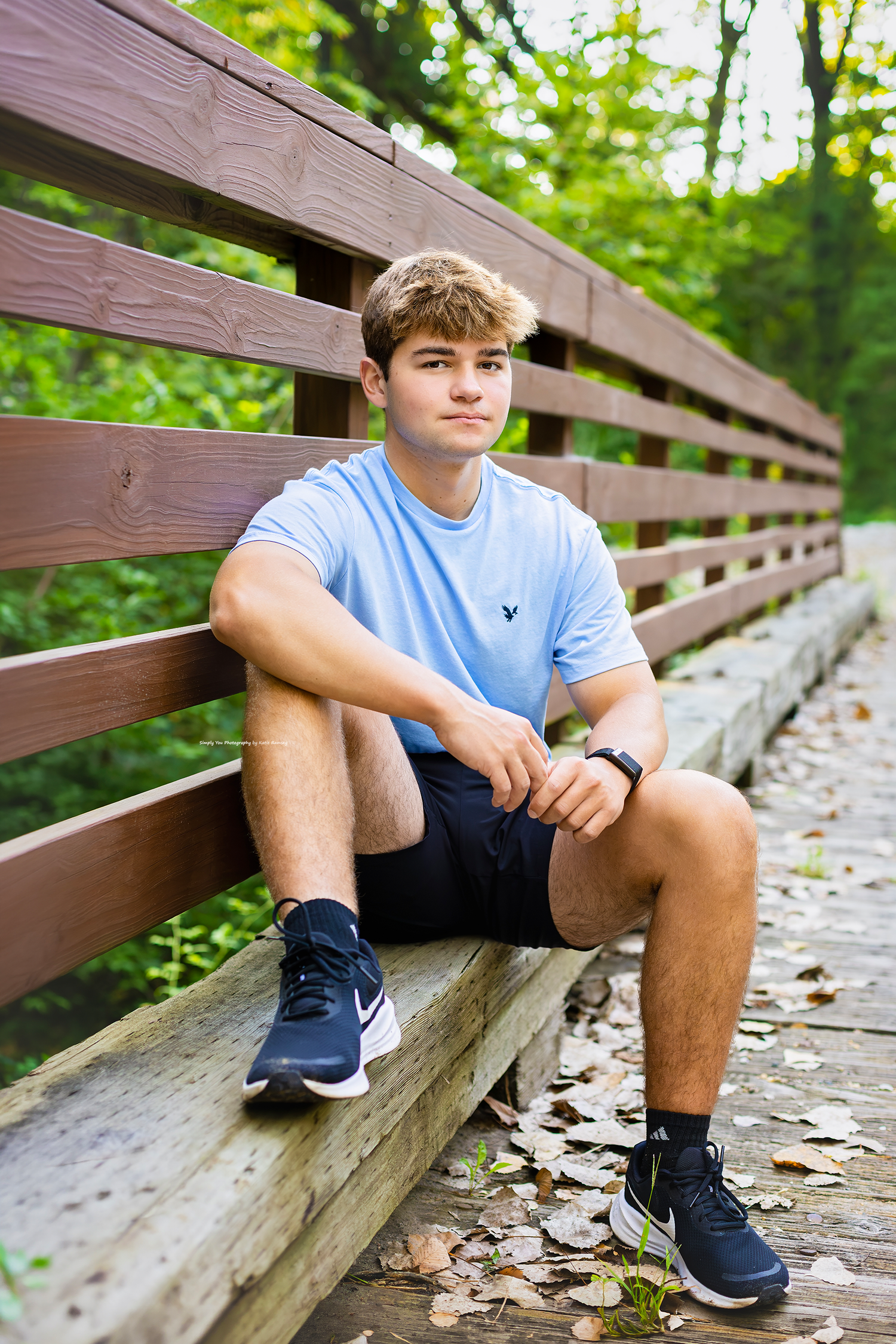 Teen boy in light blue shirt and black shorts sits relaxed on a wooden bridge surrounded by green trees.
