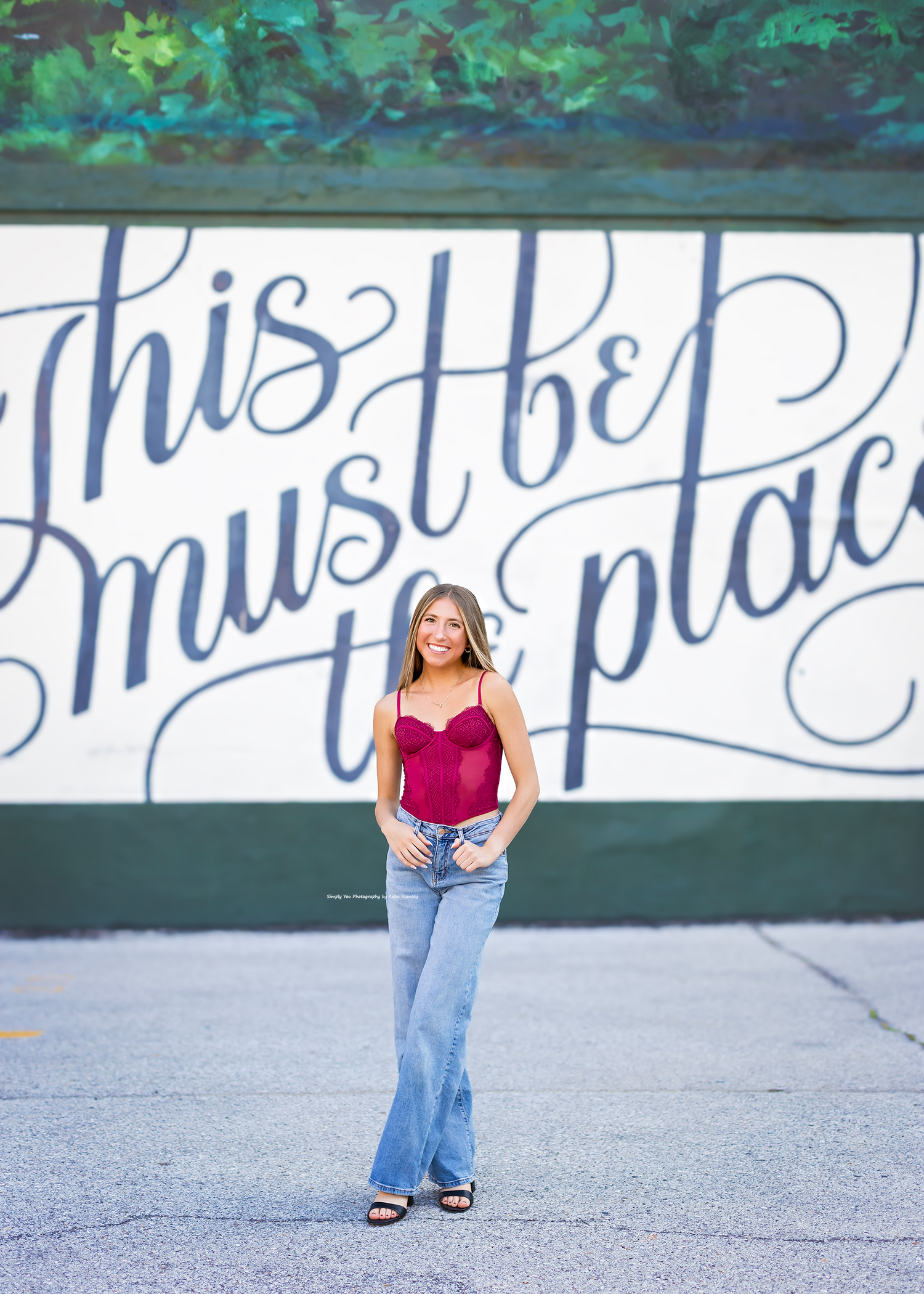 Young woman in red top and jeans posing in front of a mural reading this must be the place.