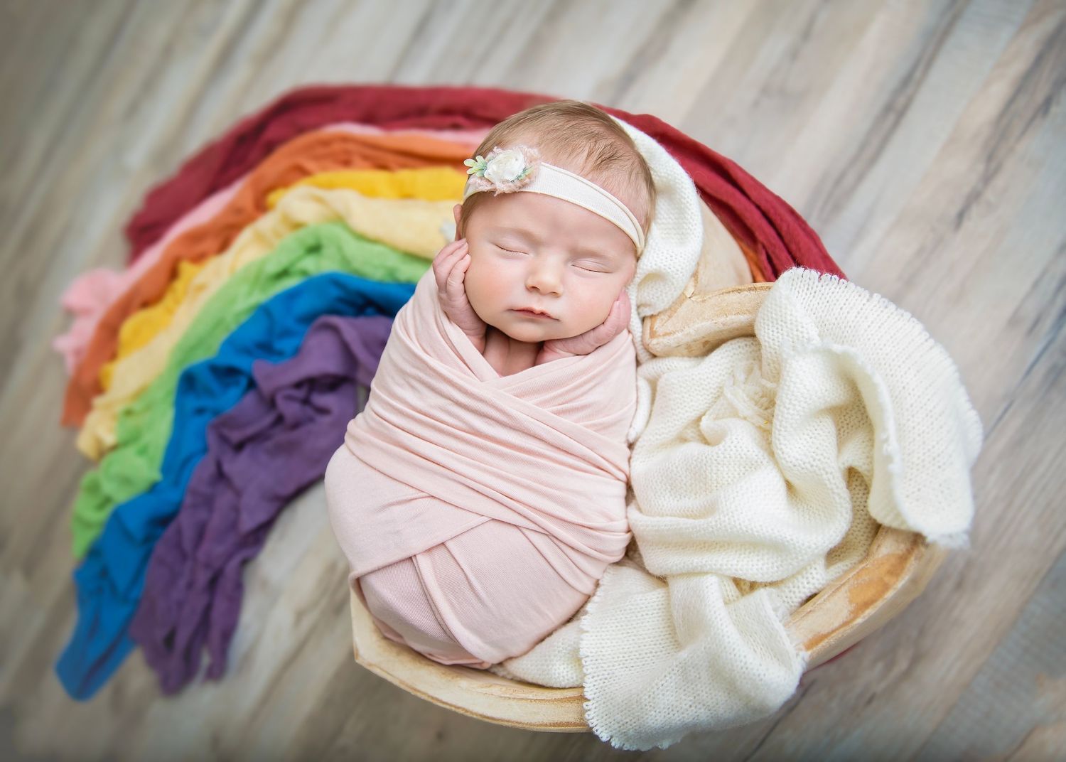 Newborn baby girl swaddled in pink wrap posed in bowl with colorful rainbow fabric backdrop.