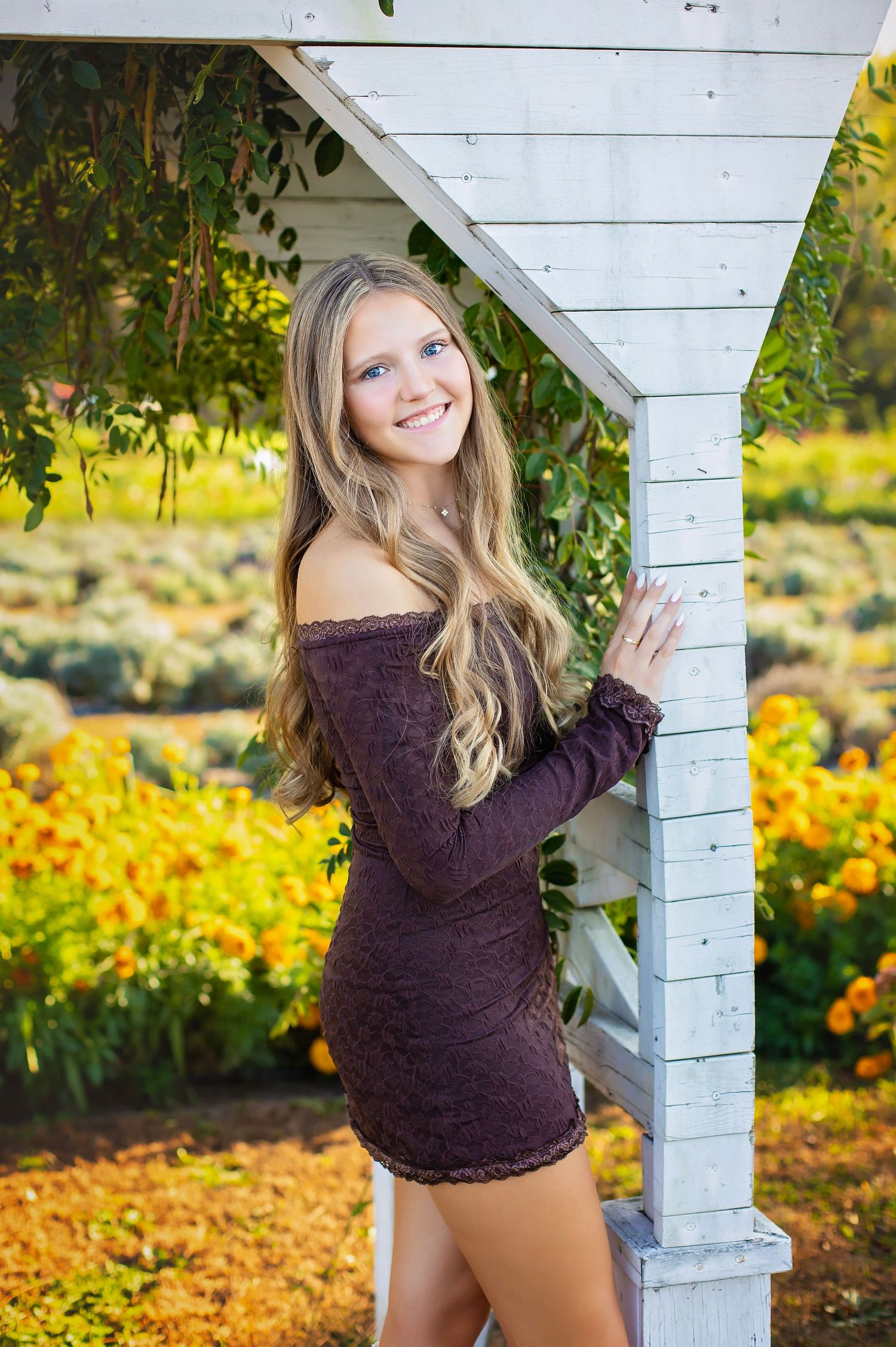 Smiling blonde teen in purple off-shoulder dress poses by white garden arbor with yellow flowers.