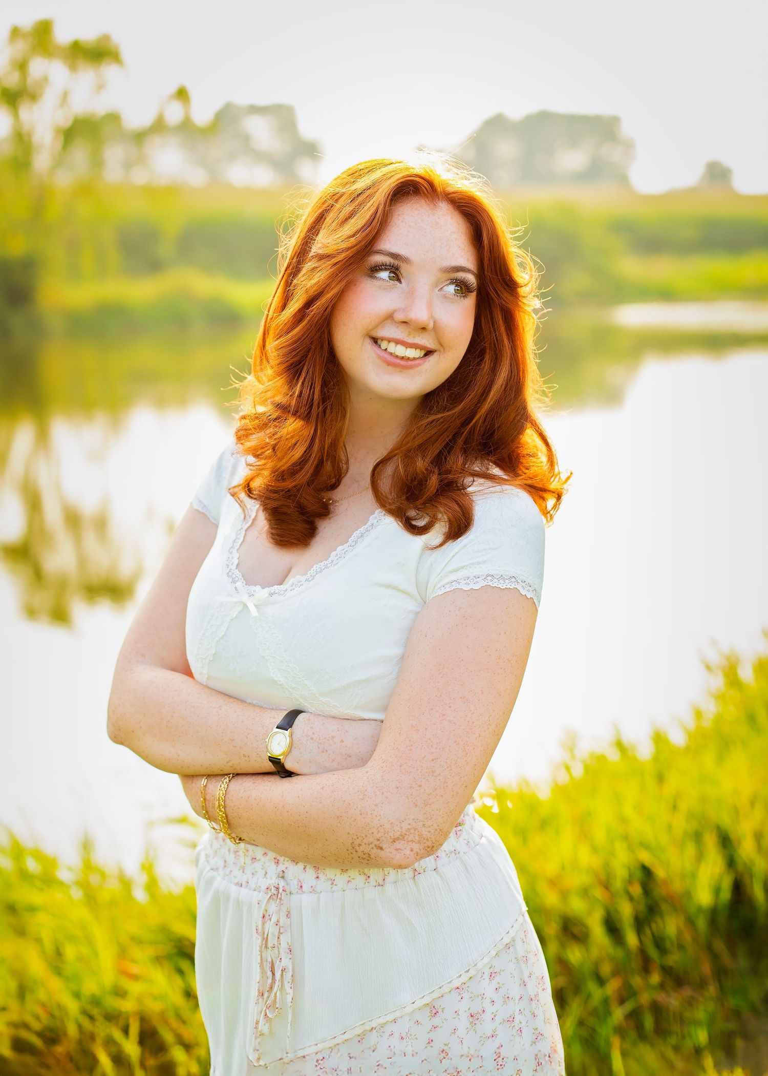 Smiling redhead teen in white dress standing by a sunlit lake with arms crossed.