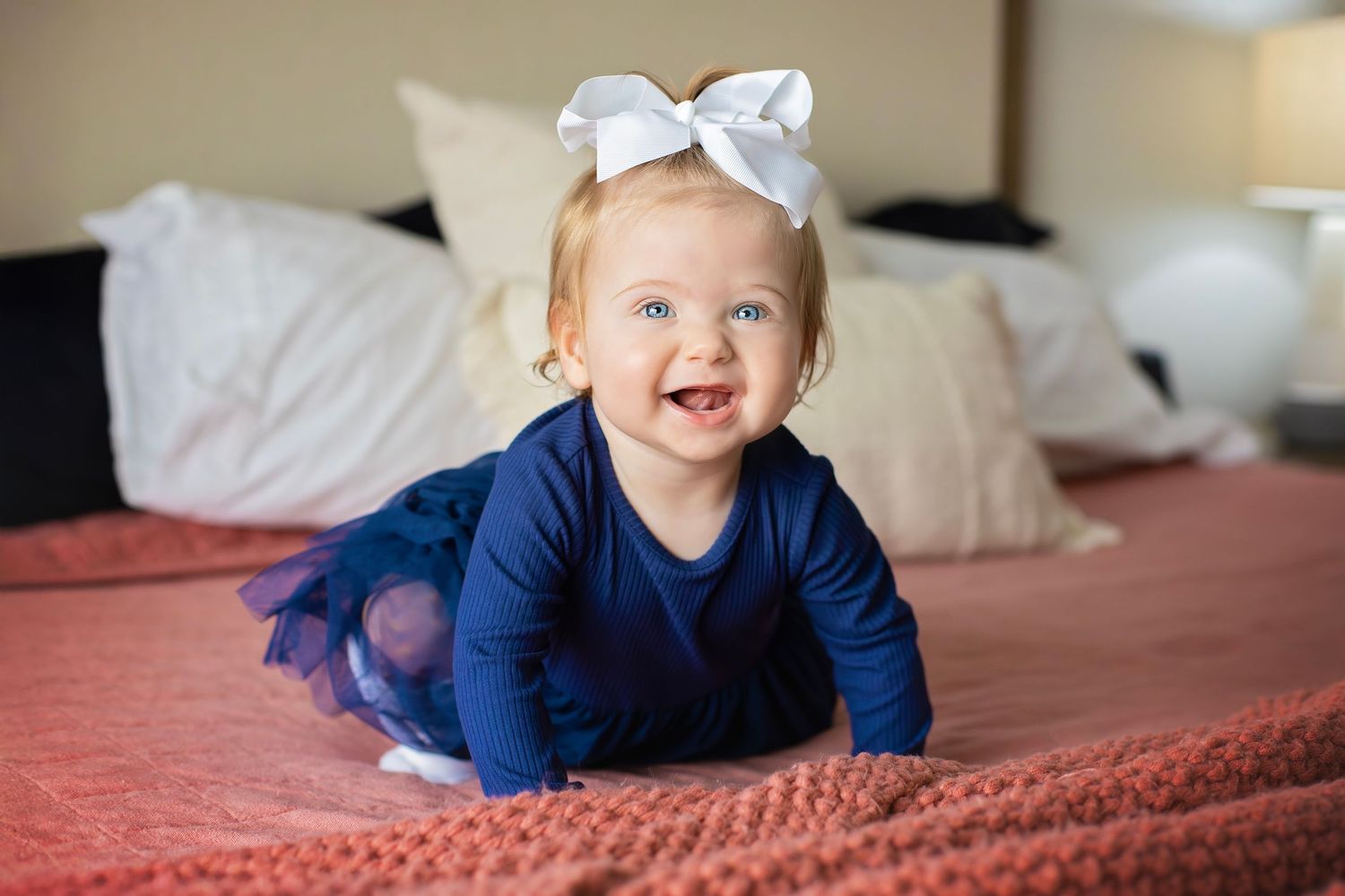 Happy baby girl in navy blue outfit with white bow crawling and smiling on a red bed.