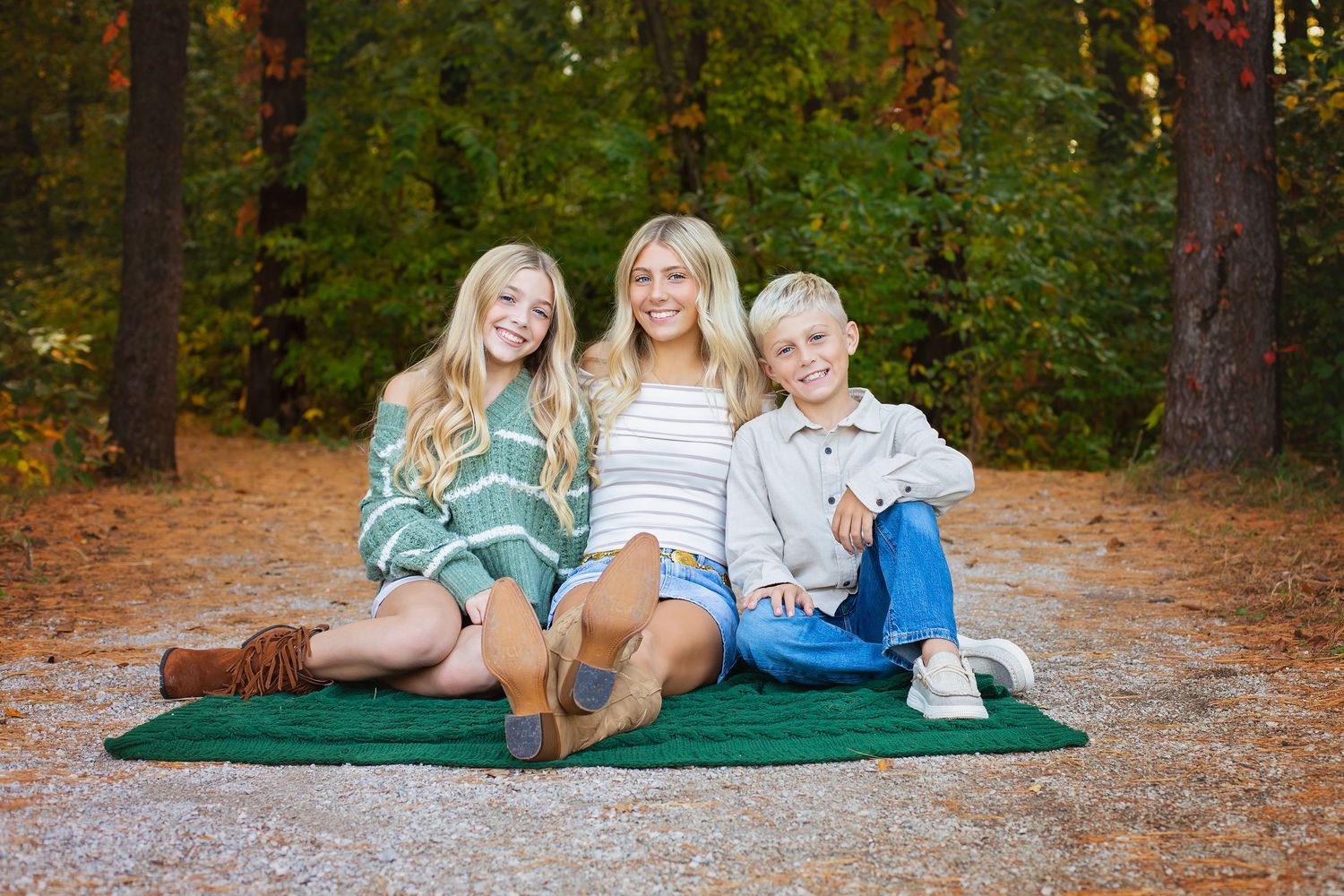 Three blonde siblings smile while sitting together on a green blanket on an autumn forest path.