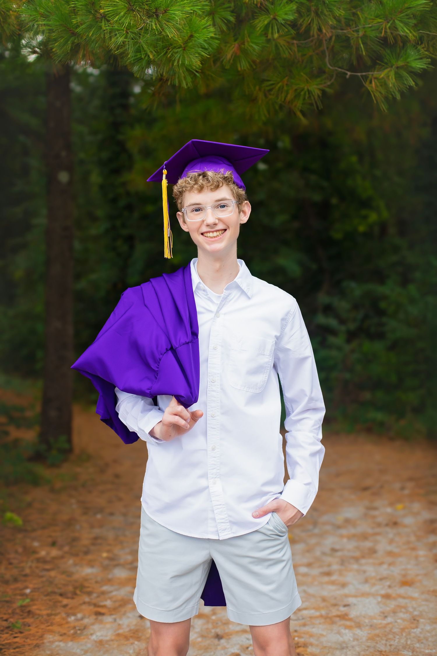 A smiling teenage boy in a white shirt holds a purple graduation cap and gown among pine trees.