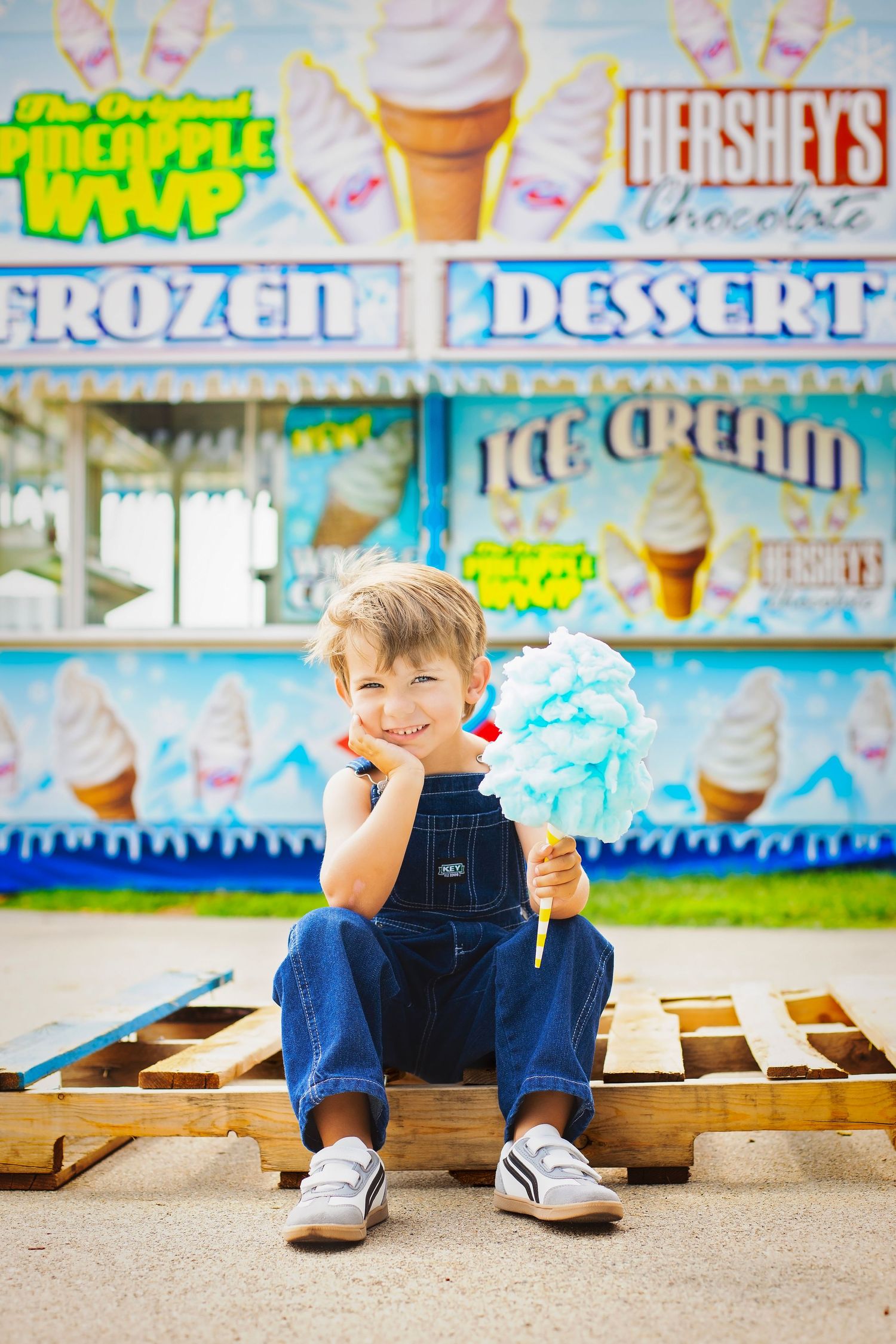 Young boy in denim overalls holding blue cotton candy at a frozen dessert fair food stand.