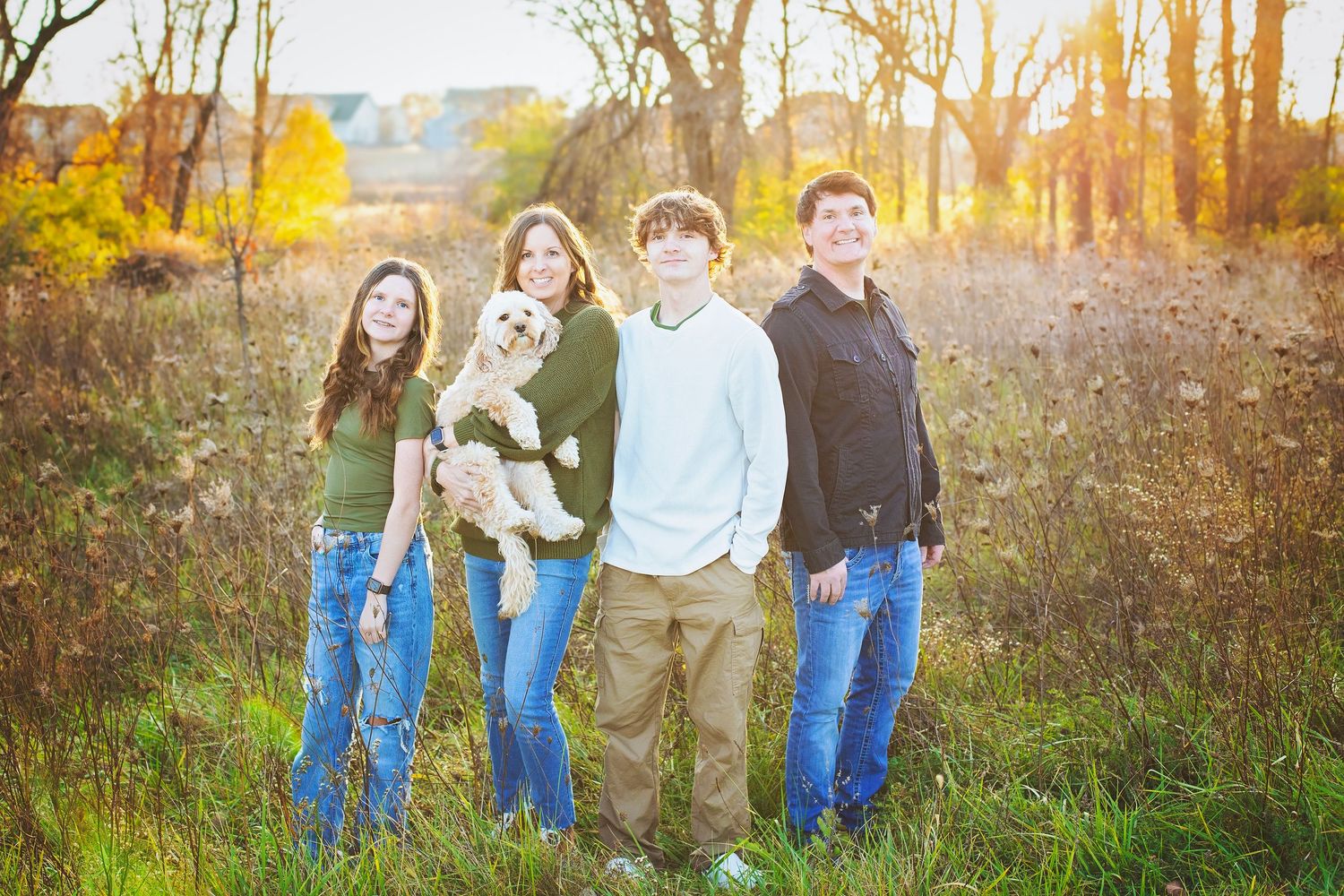 Family of four holding a puppy in a sunlit autumn meadow.