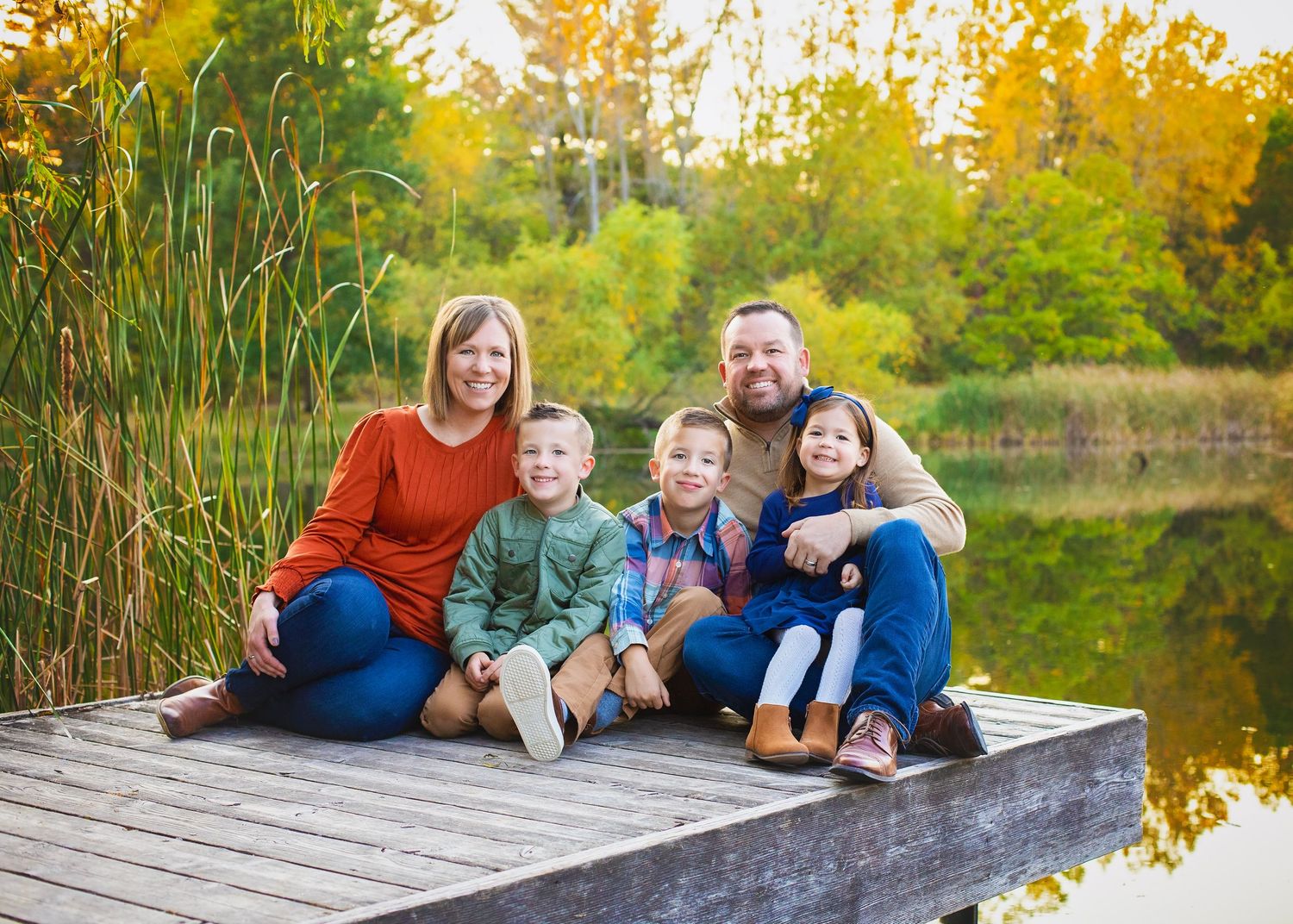 Family of five sitting on a dock by a pond in autumn foliage.