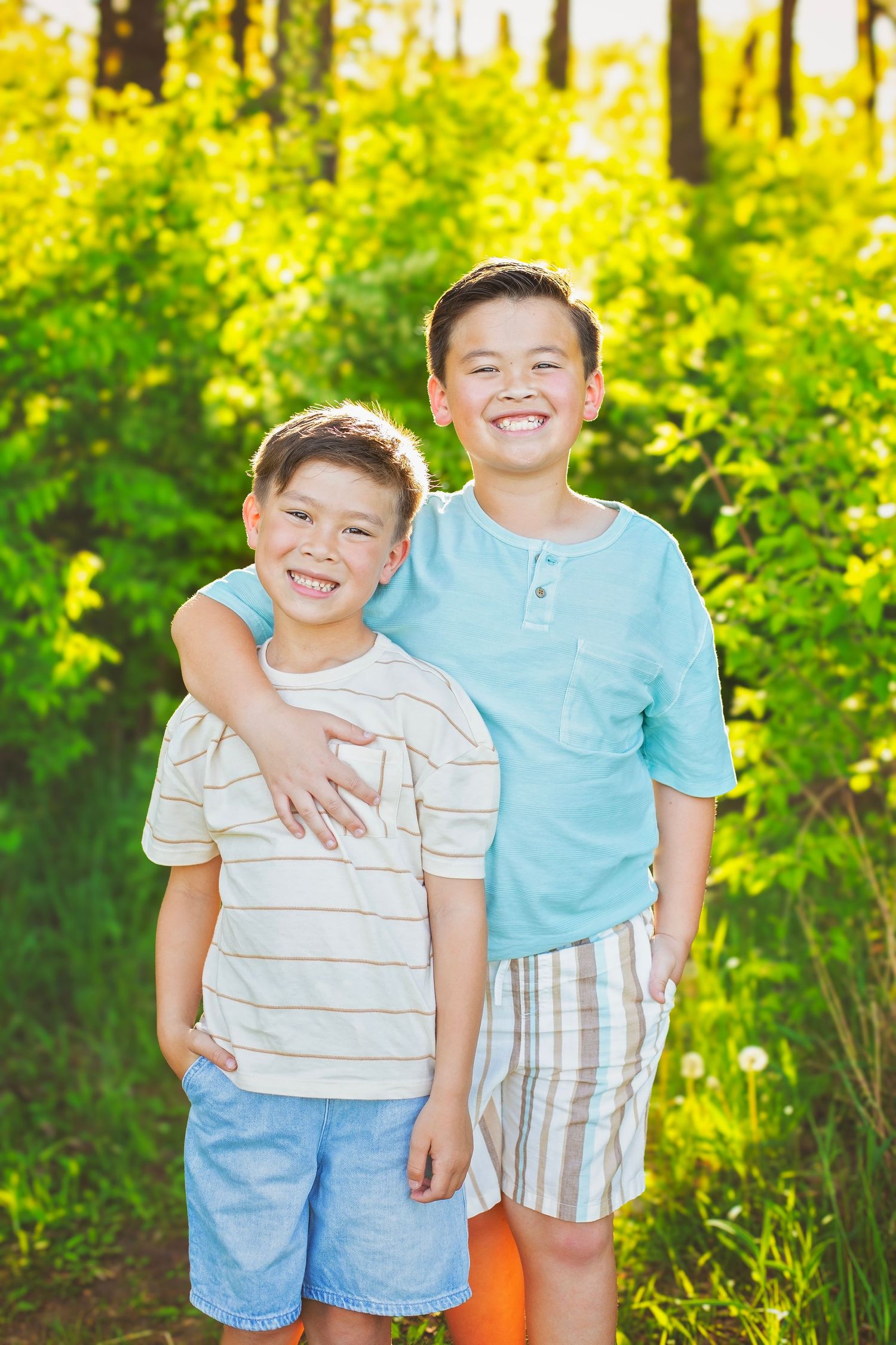 Two smiling boys pose together outdoors, the taller boy hugging the younger one amid lush green foliage.