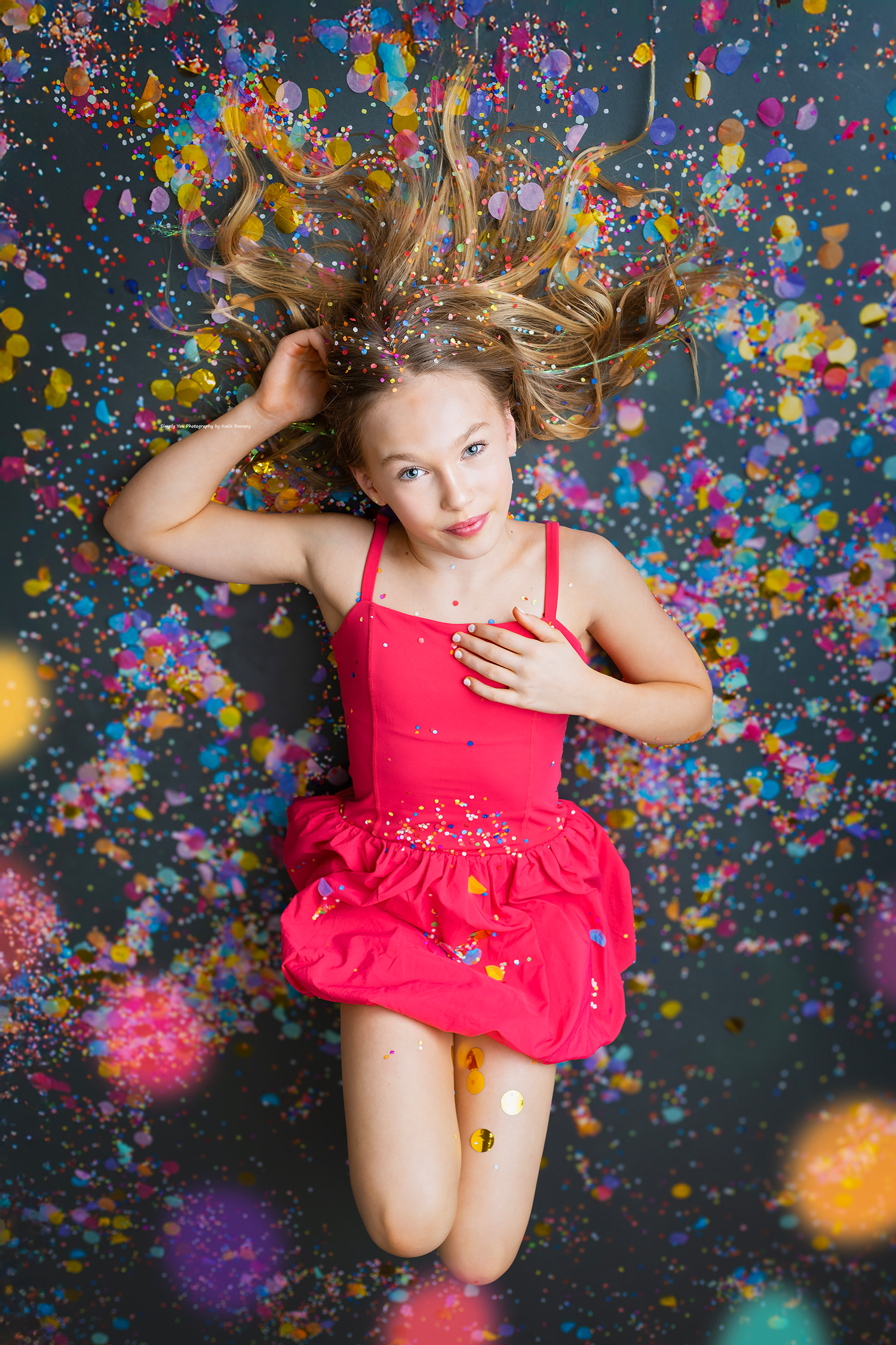 Young girl in a pink dress lying among colorful confetti with hair spread out on a dark background.
