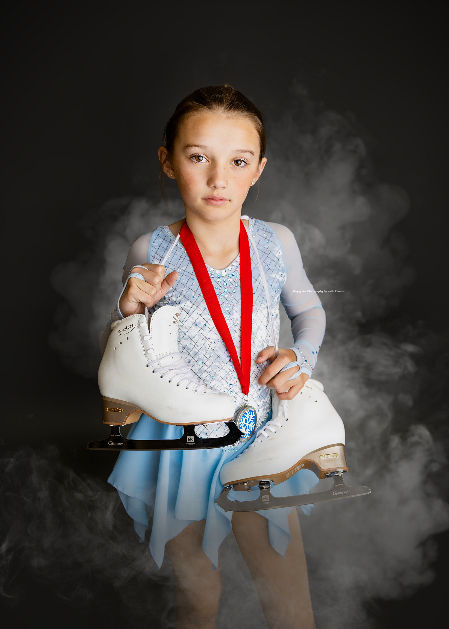 Young figure skater in blue dress holds white ice skates and wears a red ribbon medal against dark smoky background.