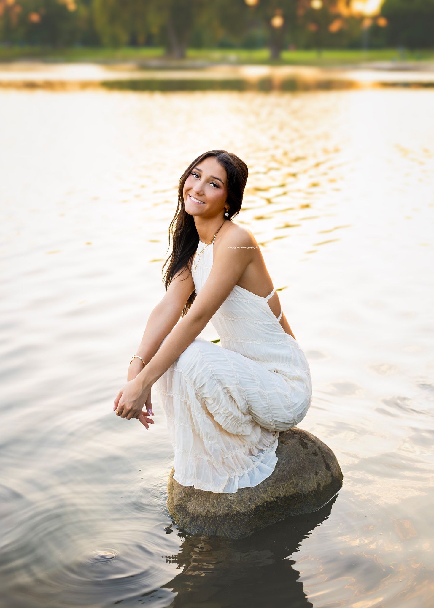 Young woman in white lace dress sitting on a rock in a lake at golden hour, smiling over her shoulder.
