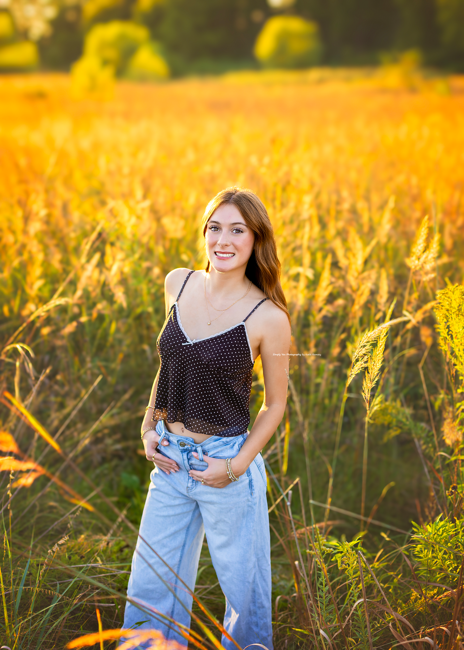 Young woman smiling in a golden field at sunset, wearing a black polka dot top and light blue jeans.