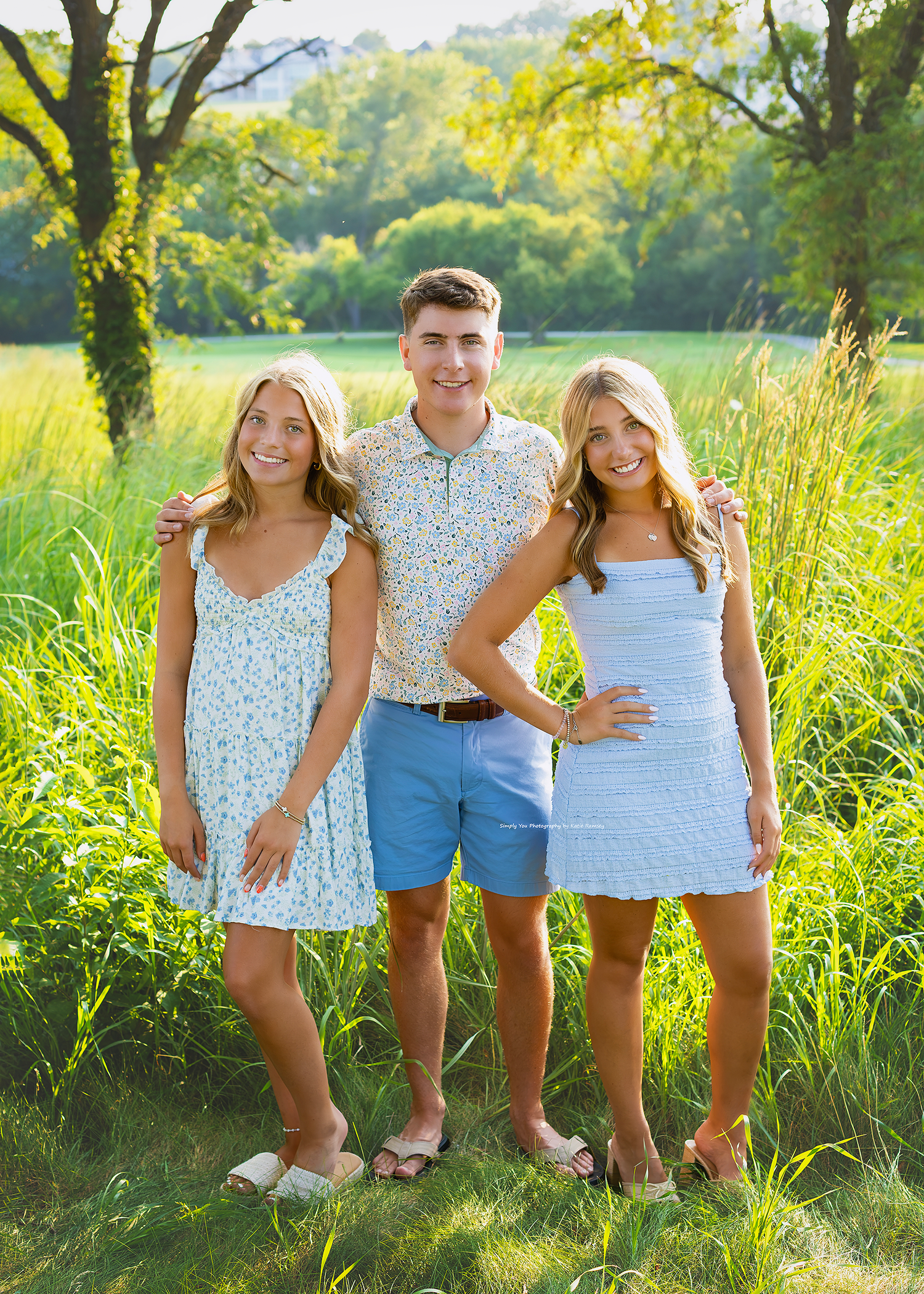 Three smiling teens pose outdoors in a sunny green field, wearing light summer outfits of blue and floral patterns.