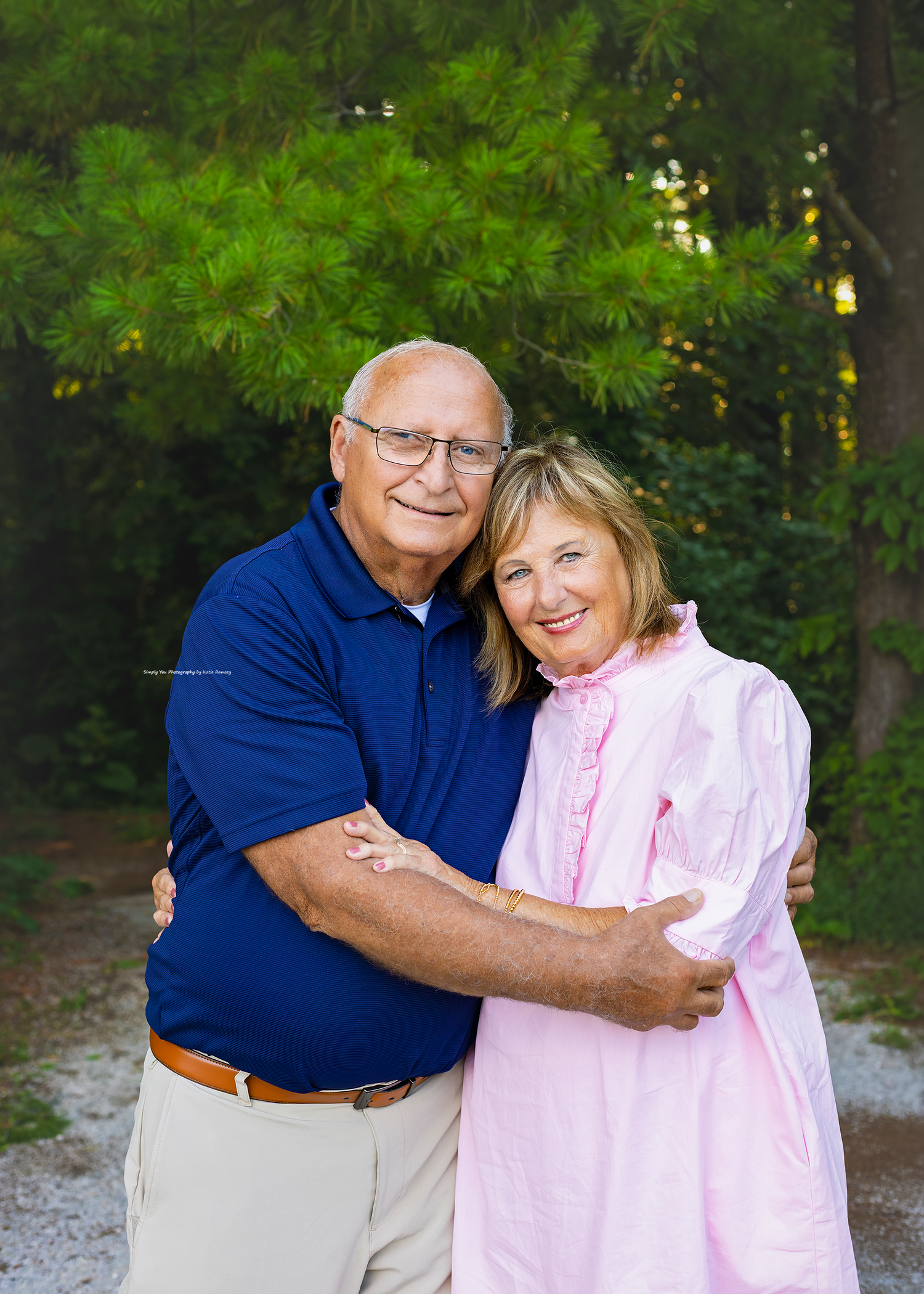 Smiling older couple embracing outdoors on a gravel path surrounded by lush green trees.