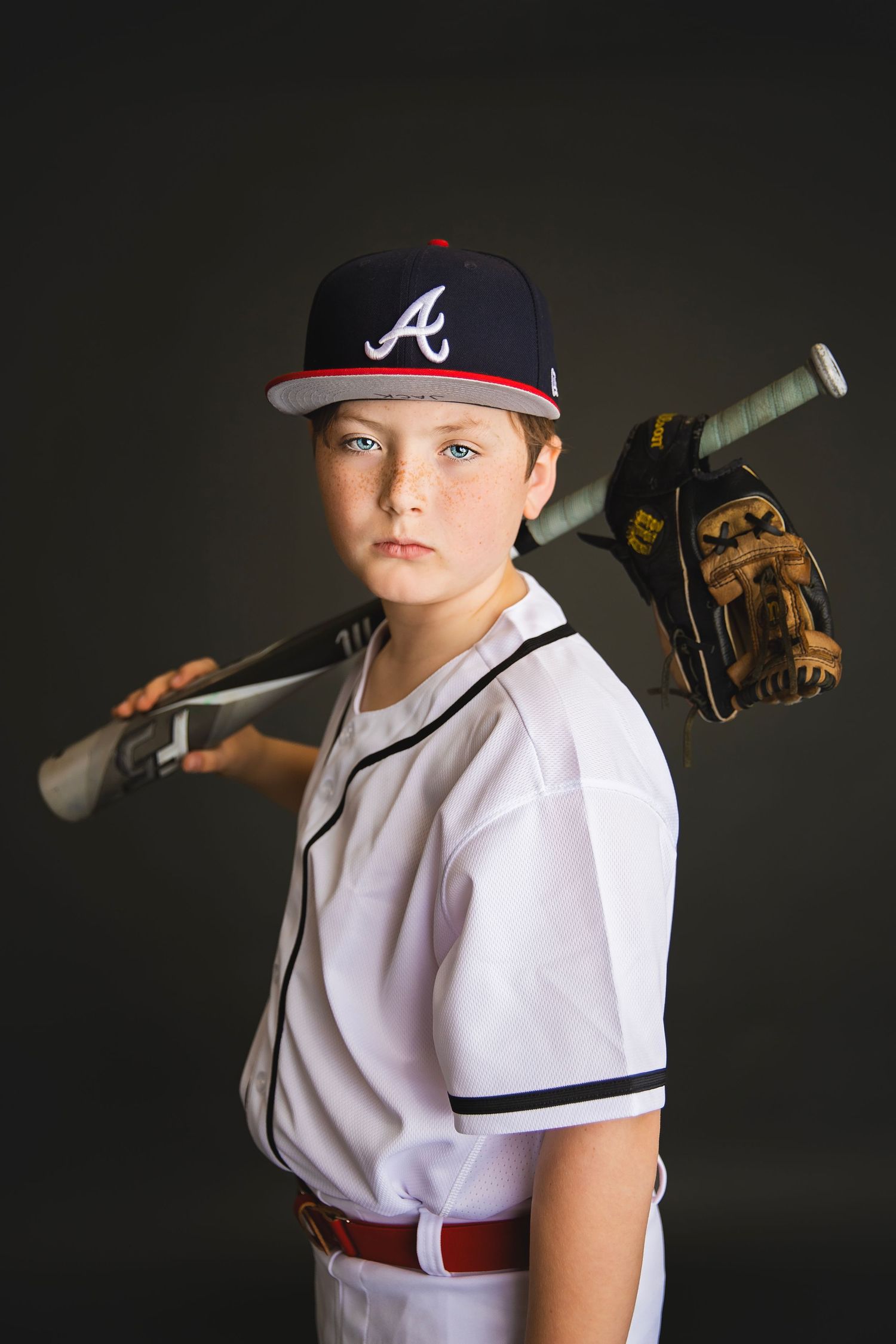 Serious young baseball player in Braves cap holding bat and glove over shoulders, dark background.