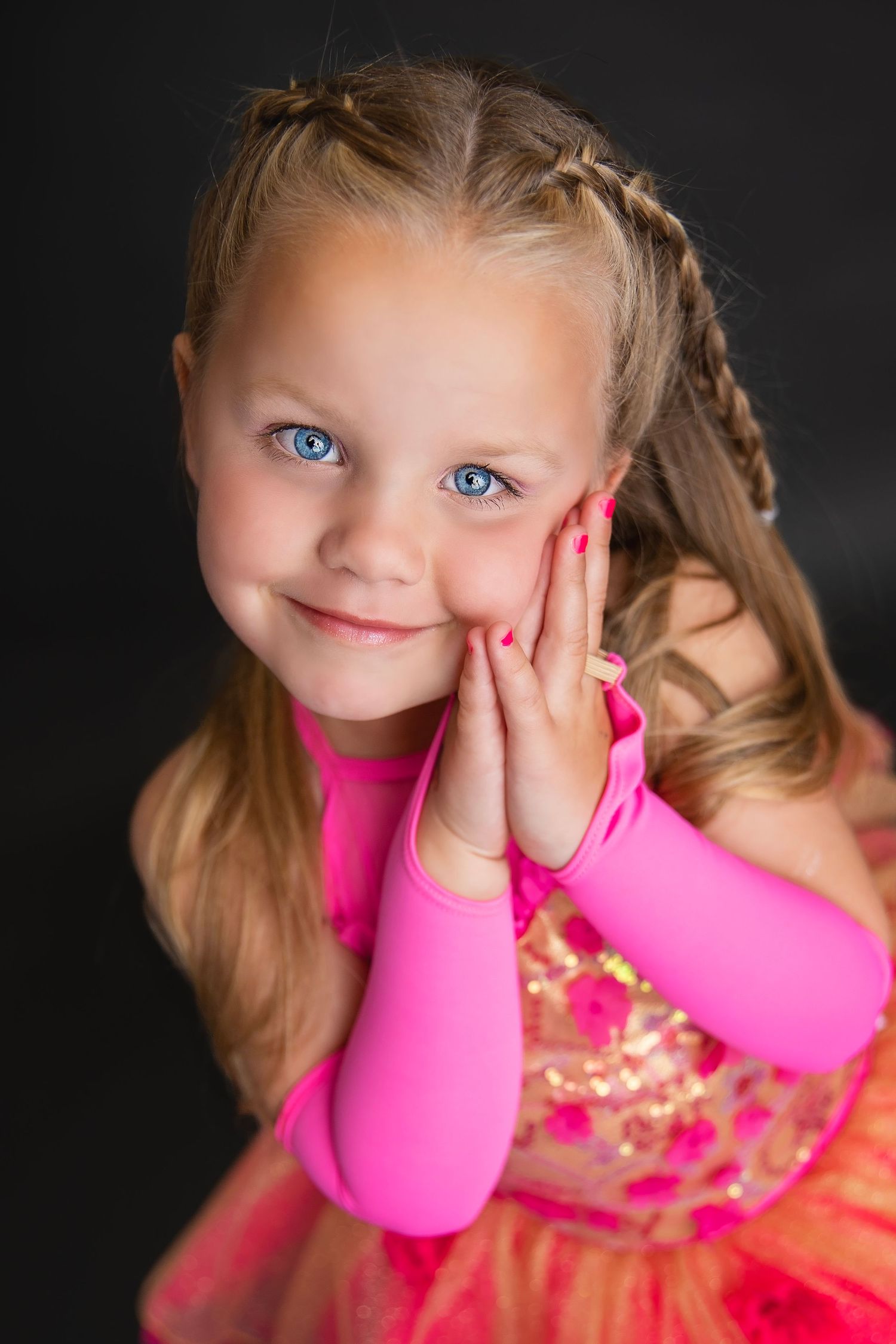 Adorable young girl with blue eyes in pink sequined dance costume smiling with hands on cheeks.