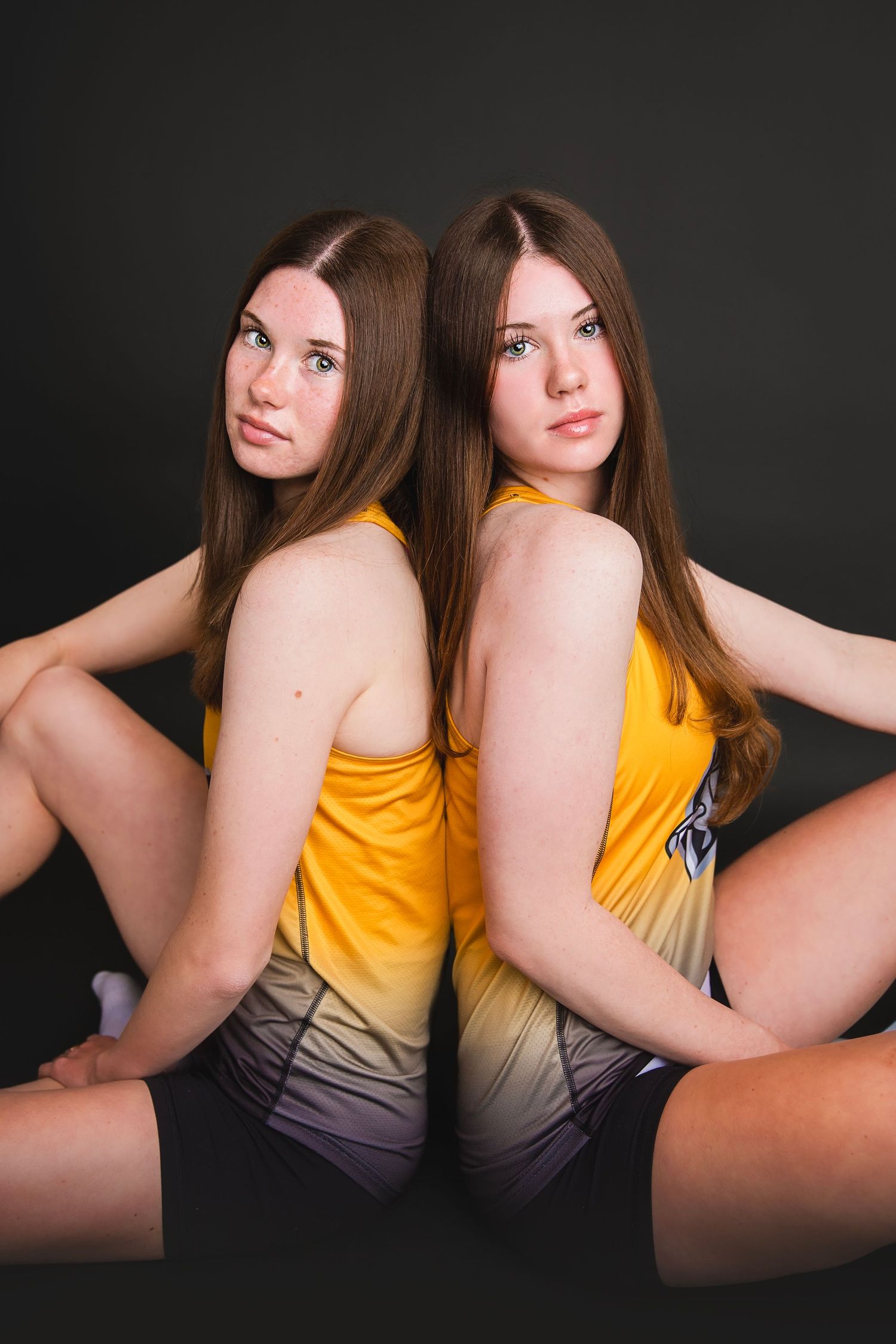 Two teenage girls in matching gold volleyball uniforms sitting back-to-back, dark studio background.