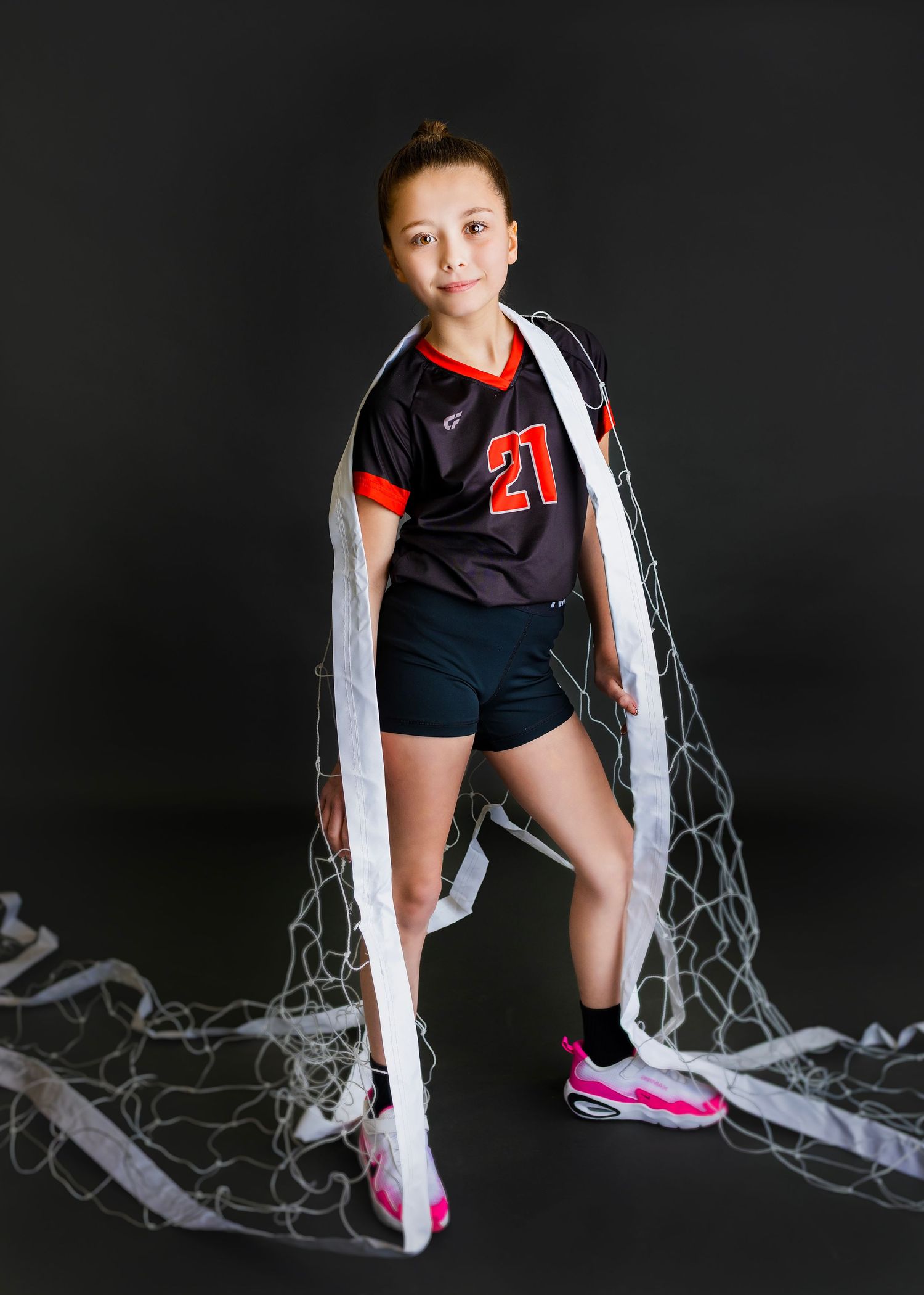 Young girl in number 21 volleyball uniform poses with a volleyball net draped around her on a dark background.