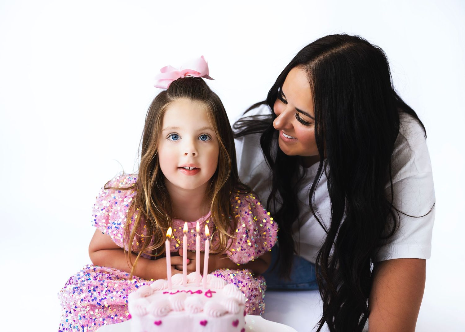 A young girl in a pink dress sits with her mom before a birthday cake with candles on a white background.