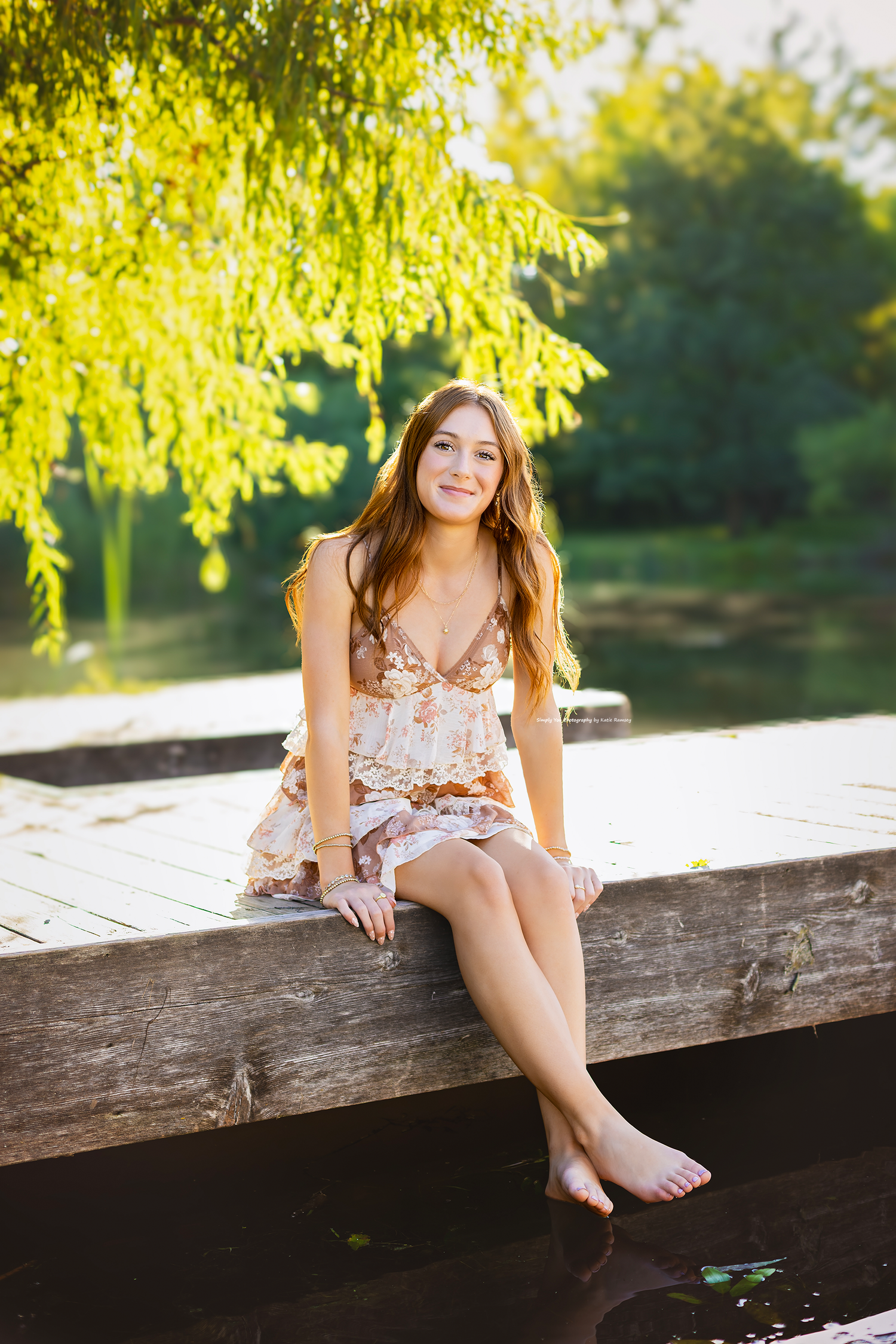 Young woman in floral dress sitting barefoot on wooden dock by pond under weeping willow in golden sunlight.