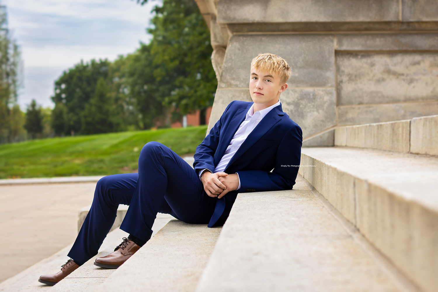 A young blonde teen boy in a navy blue suit sits on stone steps outdoors for a formal portrait photo.