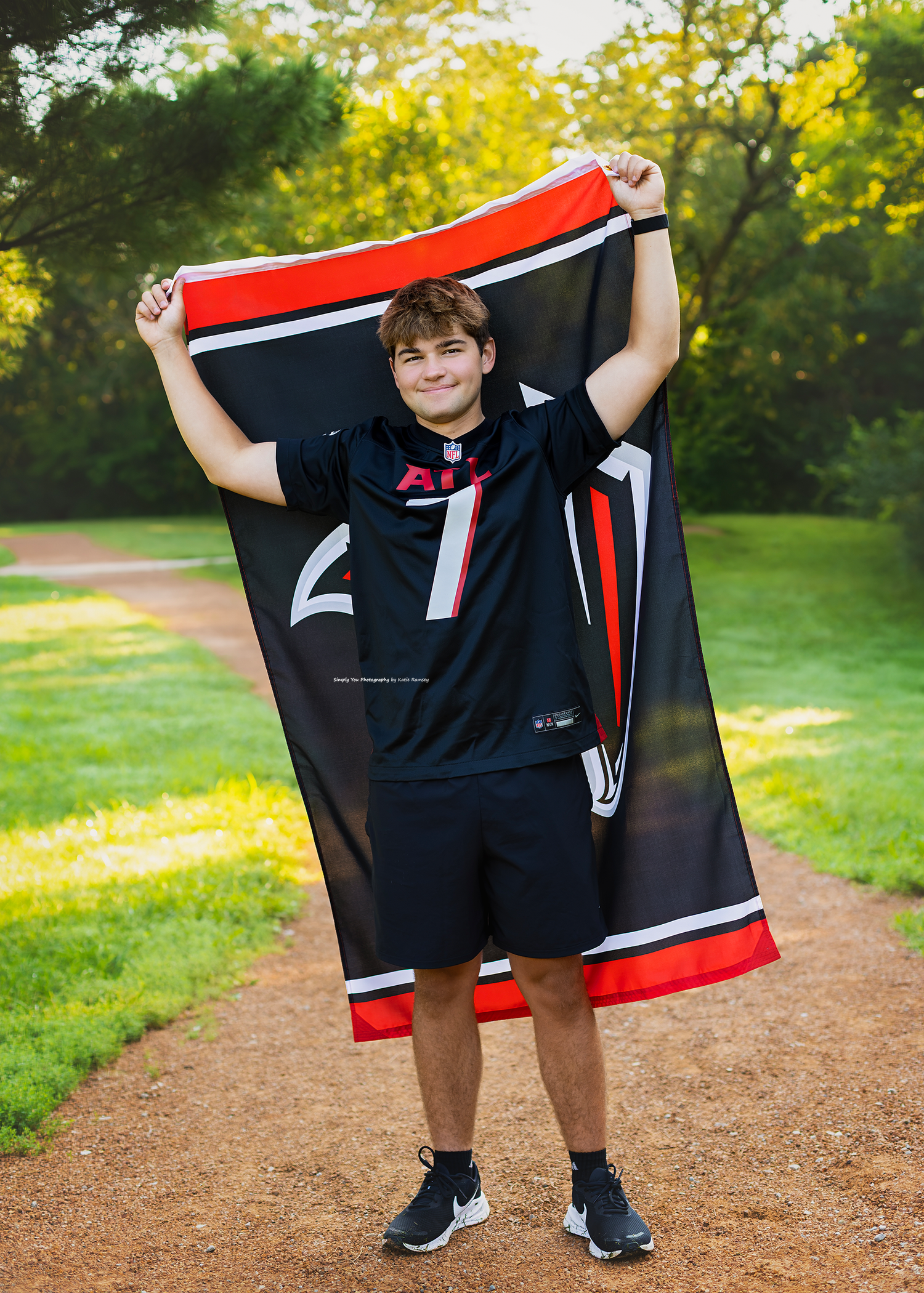 Teen boy in black NFL jersey holds up a matching flag on a sunlit park path surrounded by green trees.