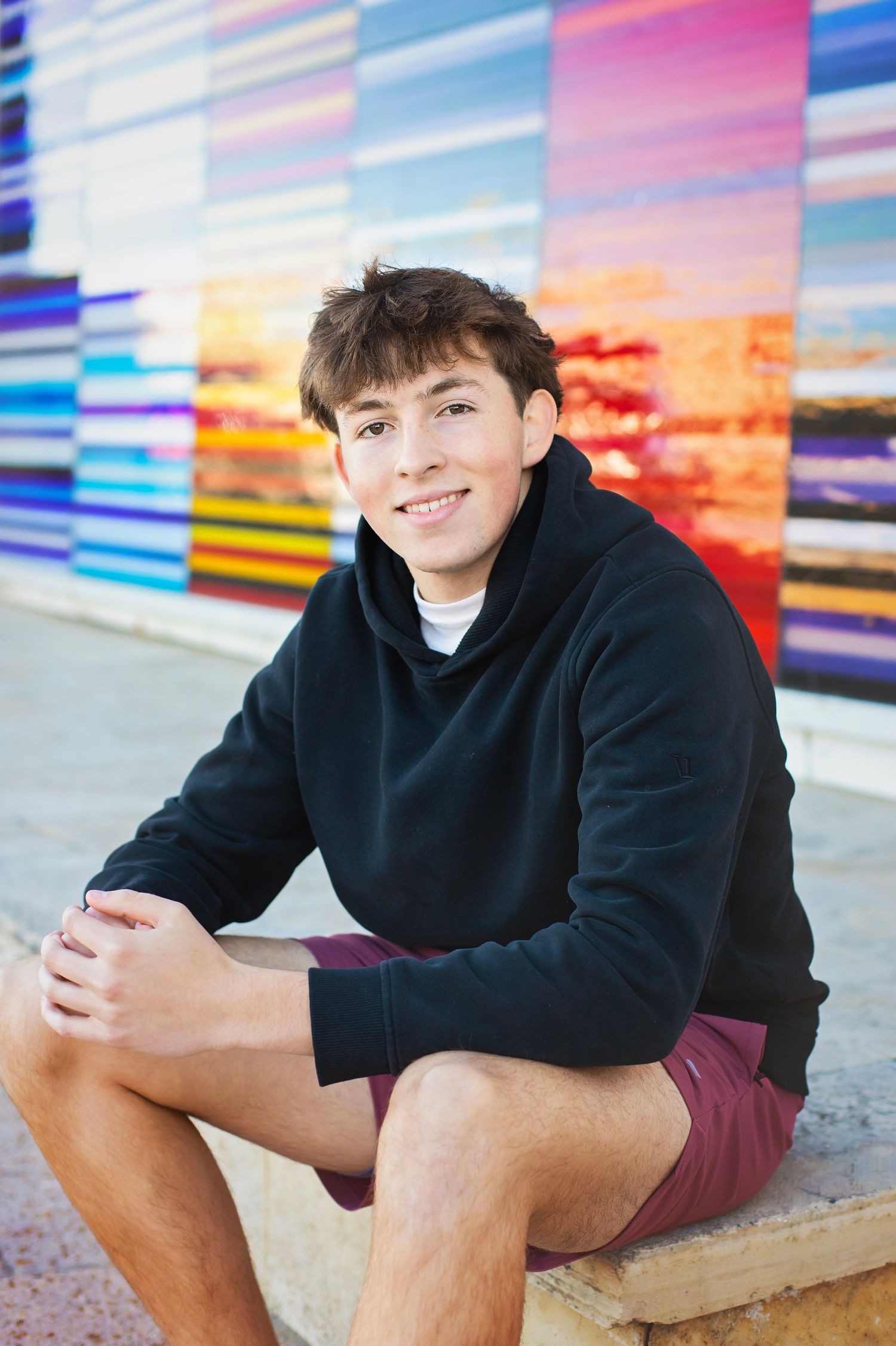 Smiling teen boy in black hoodie sits before vibrant colorful mural wall.