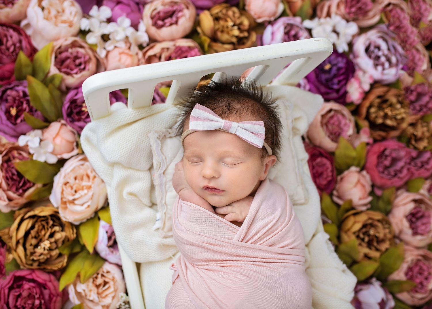 Sleeping newborn baby girl in pink swaddle with white bow headband surrounded by colorful flowers.