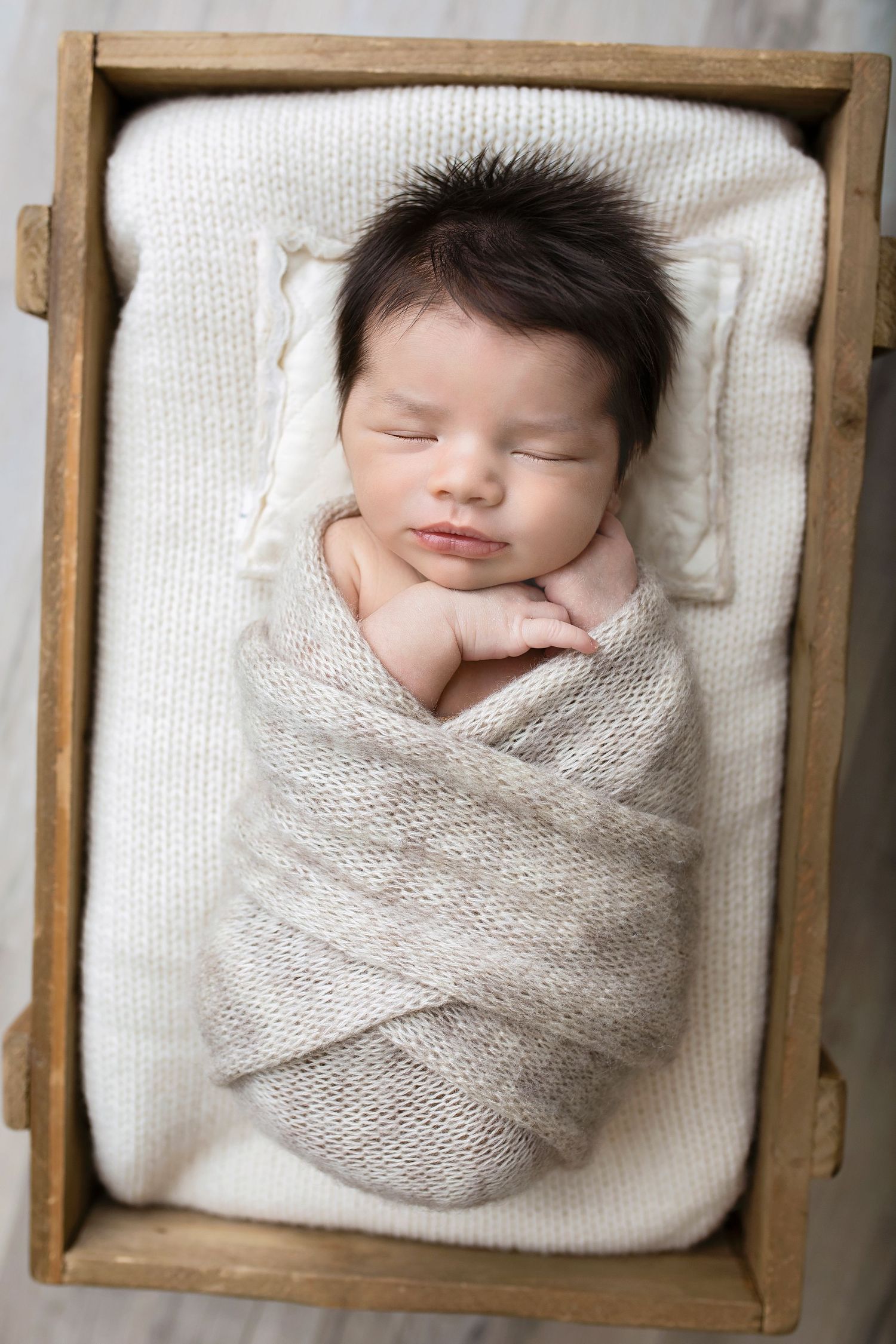 Sleeping newborn baby wrapped in beige knit swaddle resting in a rustic wooden crate with white blanket.