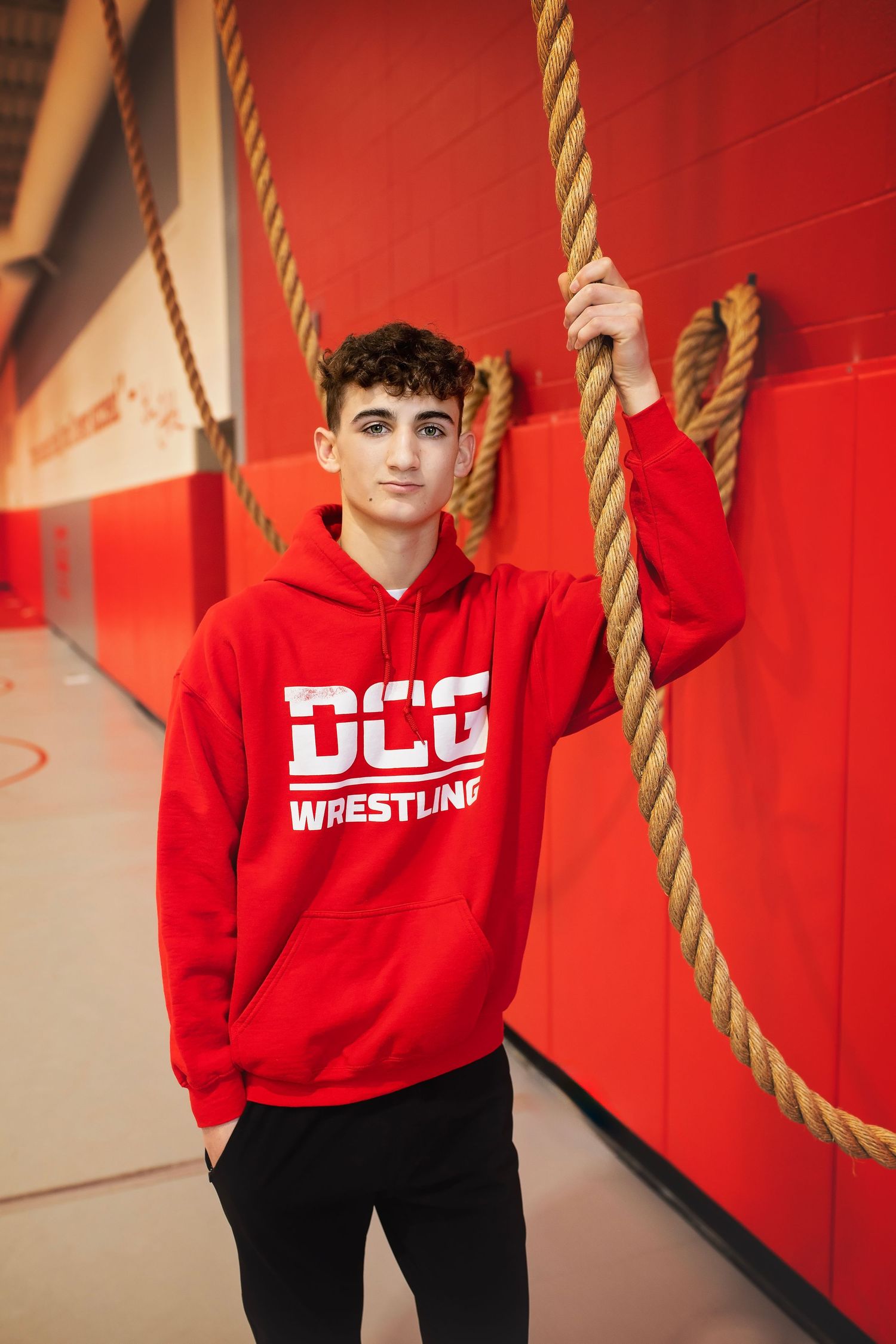 Teen in red DCG Wrestling hoodie leaning against gym wall holding a thick rope.