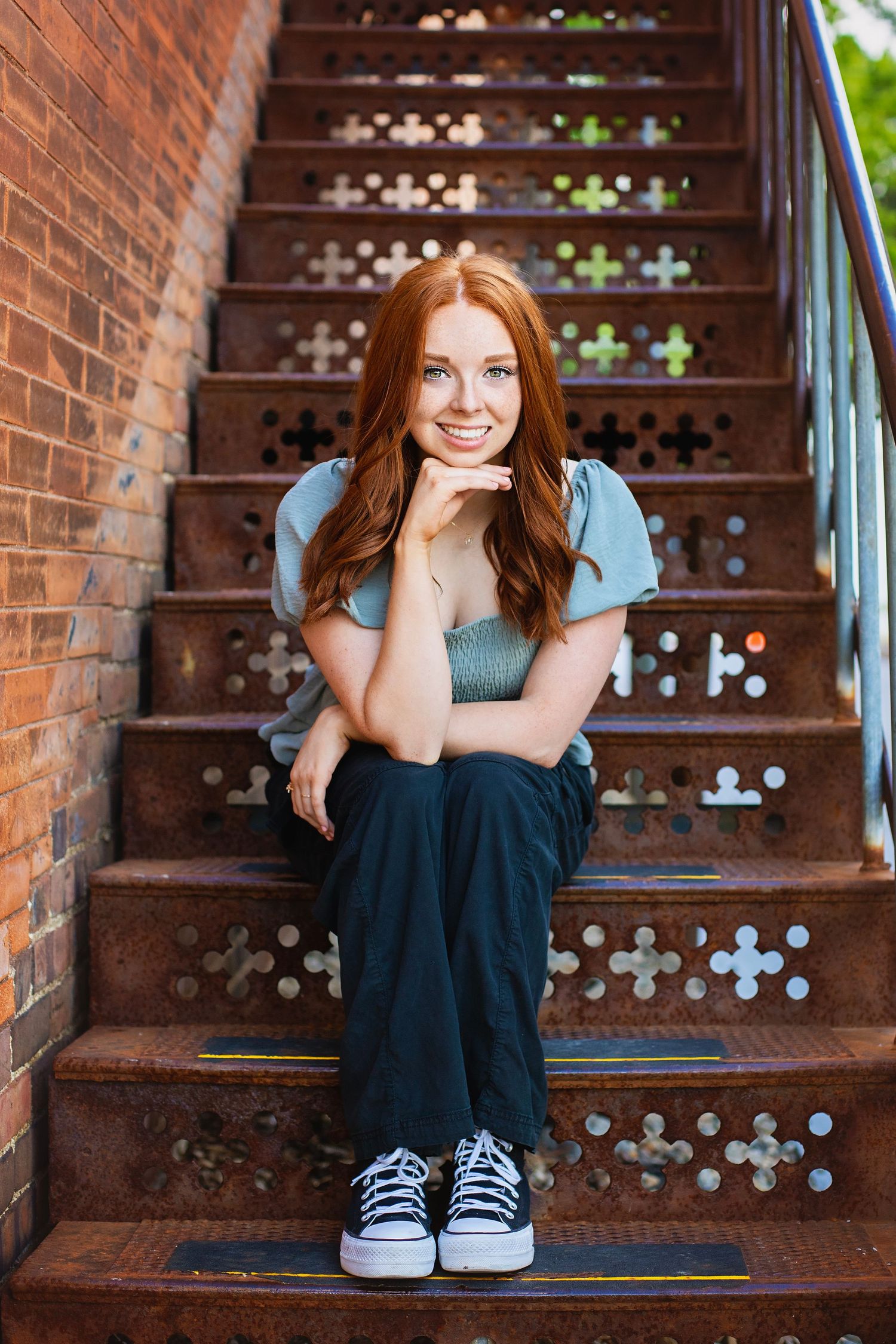 Smiling redhead teenage girl sitting on decorative metal stairs wearing casual blue outfit and sneakers.