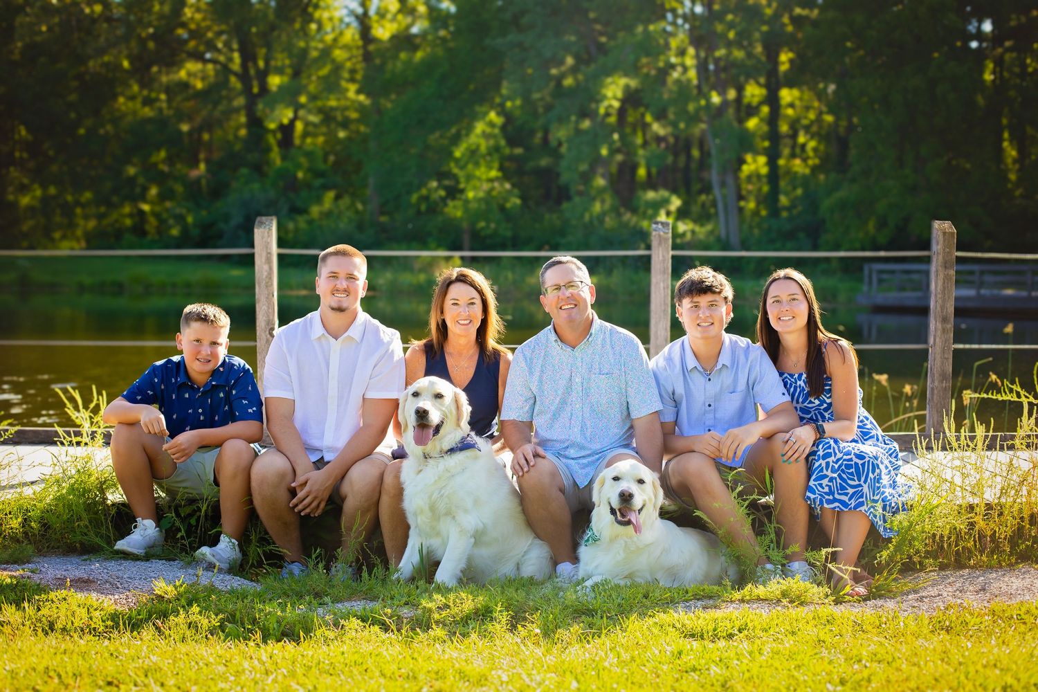 Family of six sitting outdoors with two white dogs near a pond in a sunny green landscape.