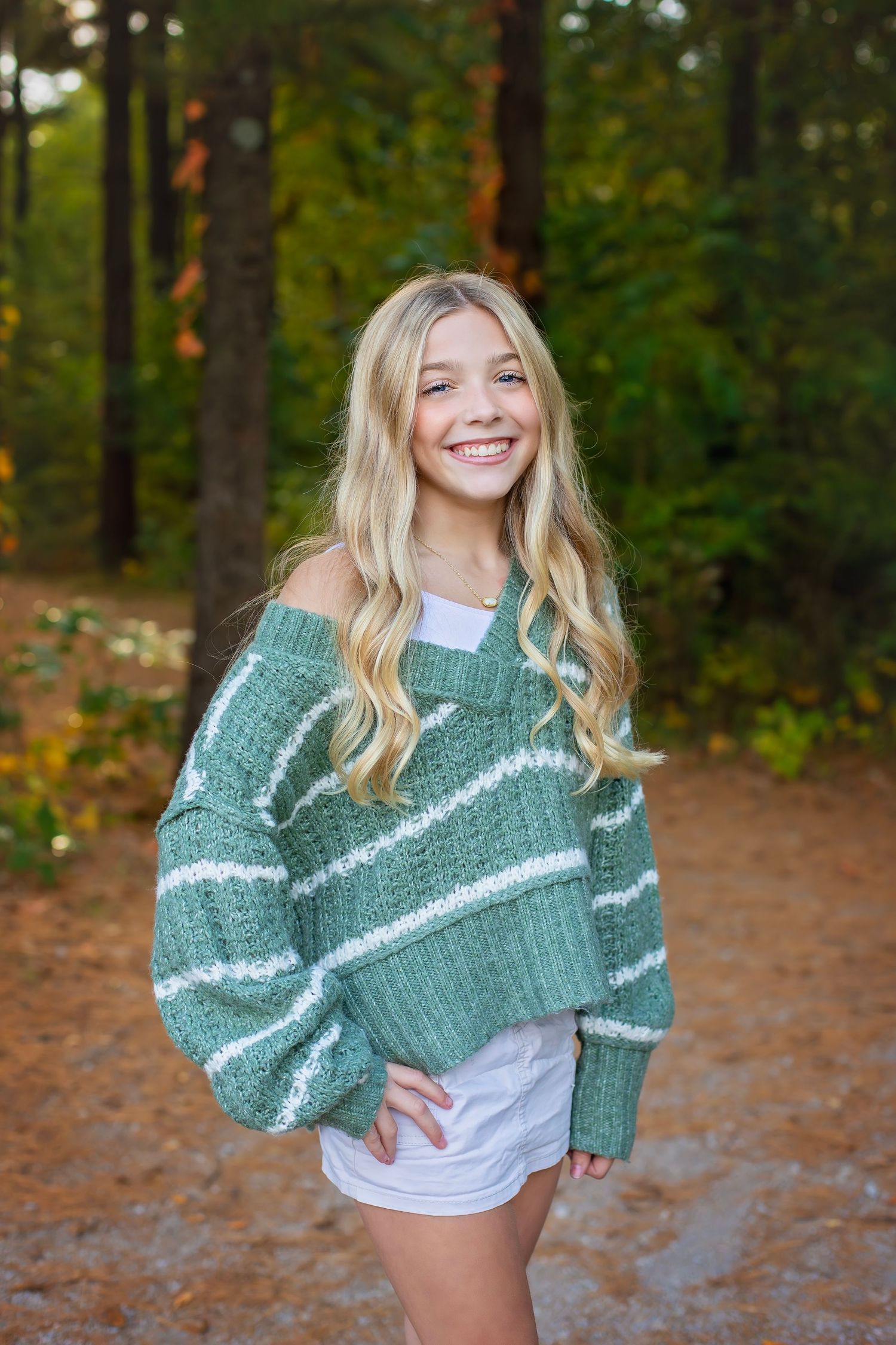 A smiling blonde girl in a green striped sweater and white shorts stands on an autumn forest path.