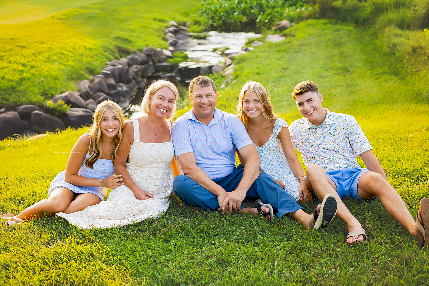 Happy family of five sitting together on green grass with a scenic waterfall backdrop outdoors.
