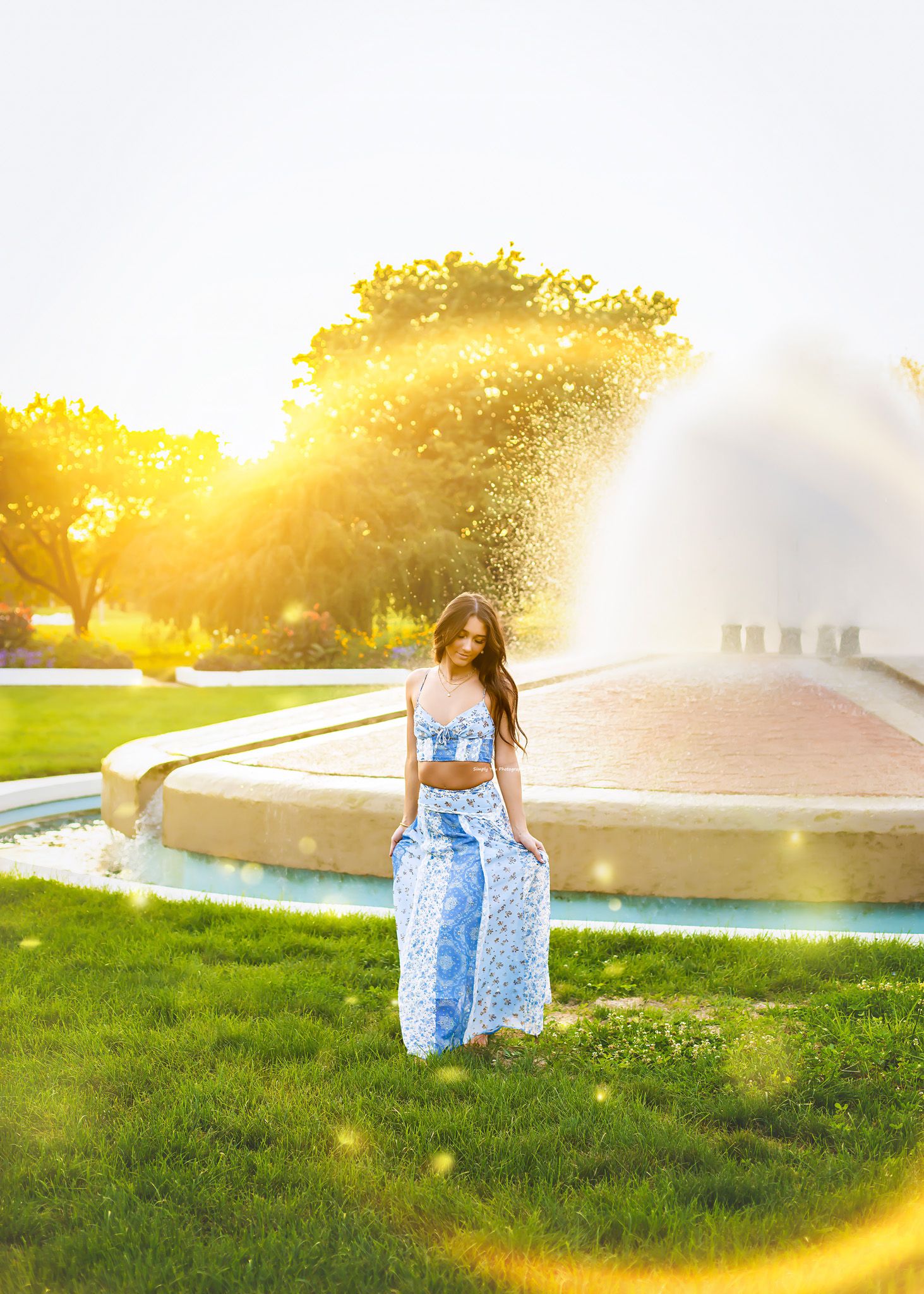Woman in blue floral two-piece outfit standing by a sunlit fountain in a lush green park.