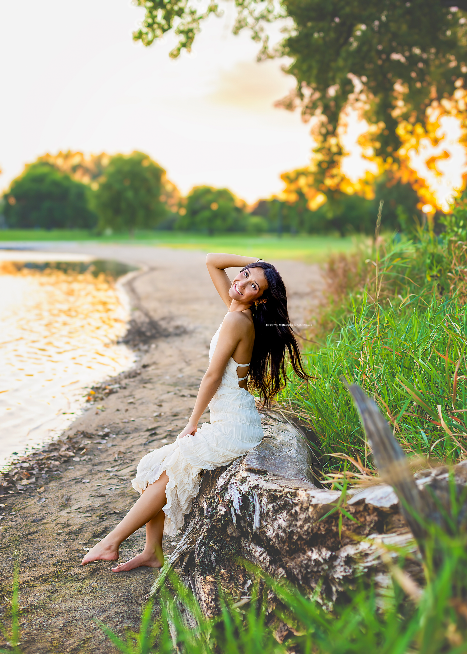 Young woman in white dress sitting on driftwood by a serene lakeside at golden sunset.