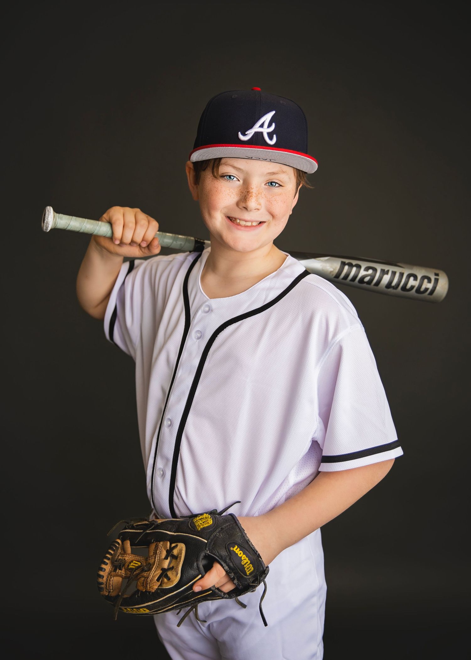 Smiling youth baseball player in Braves uniform holding Marucci bat over shoulder with glove, dark background.