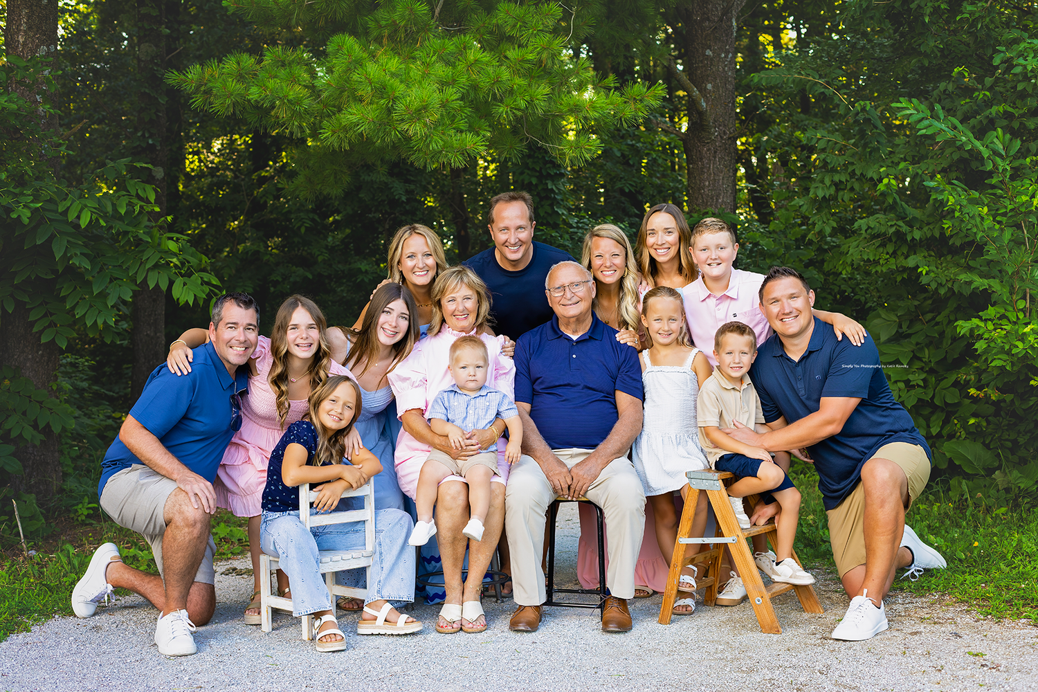Large multigenerational family portrait outdoors, smiling group of adults and children posed together near lush green trees.