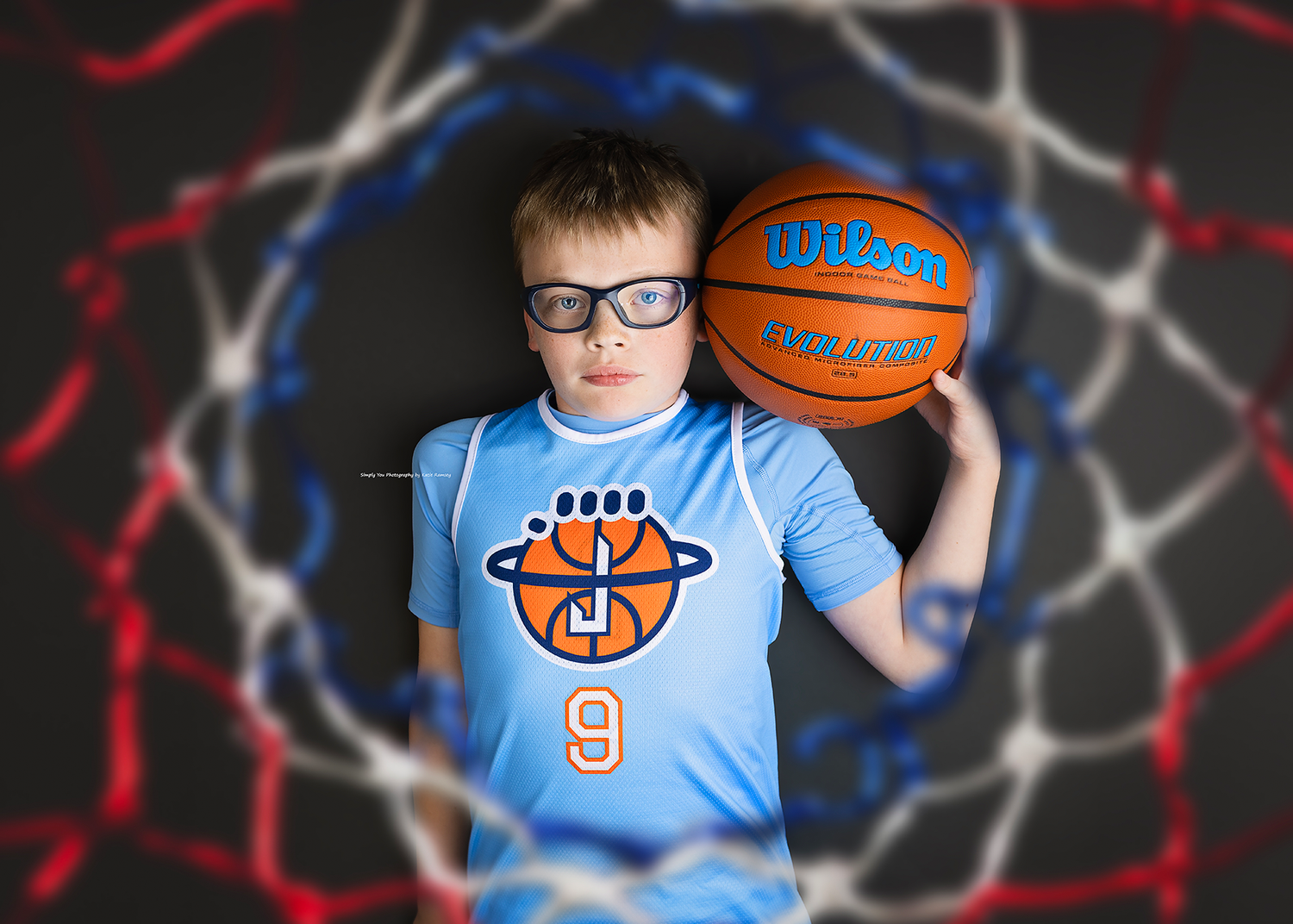 Young boy in glasses holds a Wilson basketball wearing a blue number 9 jersey in front of a net.