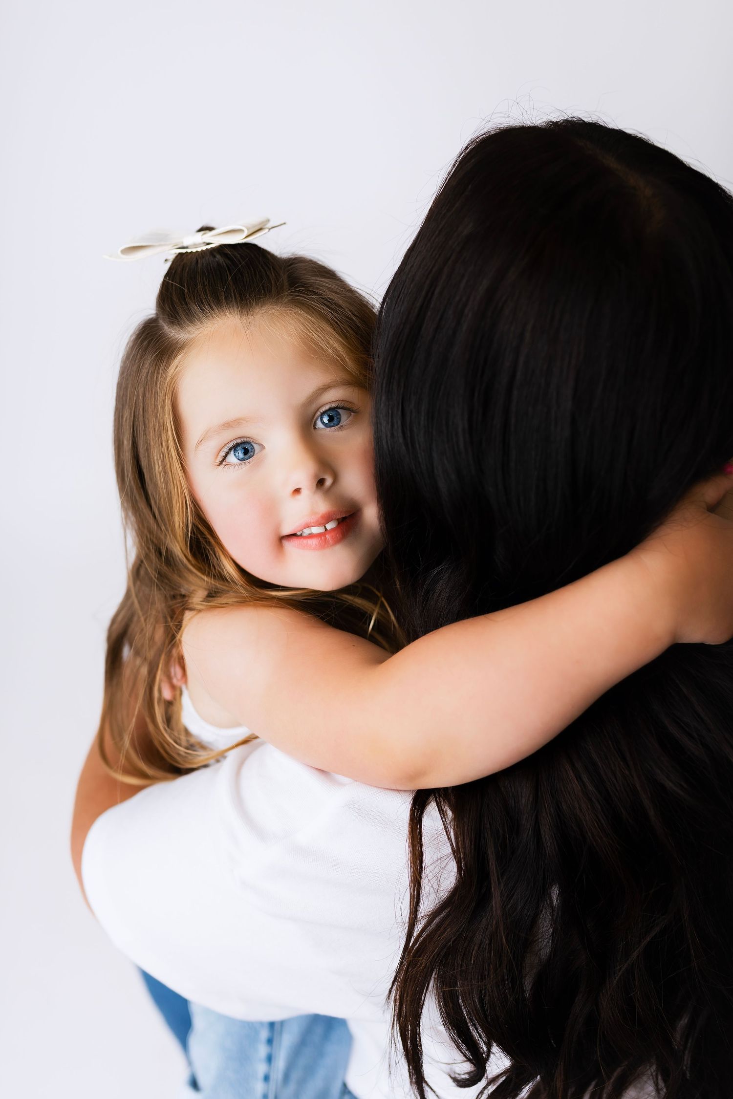 A young girl with blue eyes and a bow hugs a dark-haired woman against a white background.