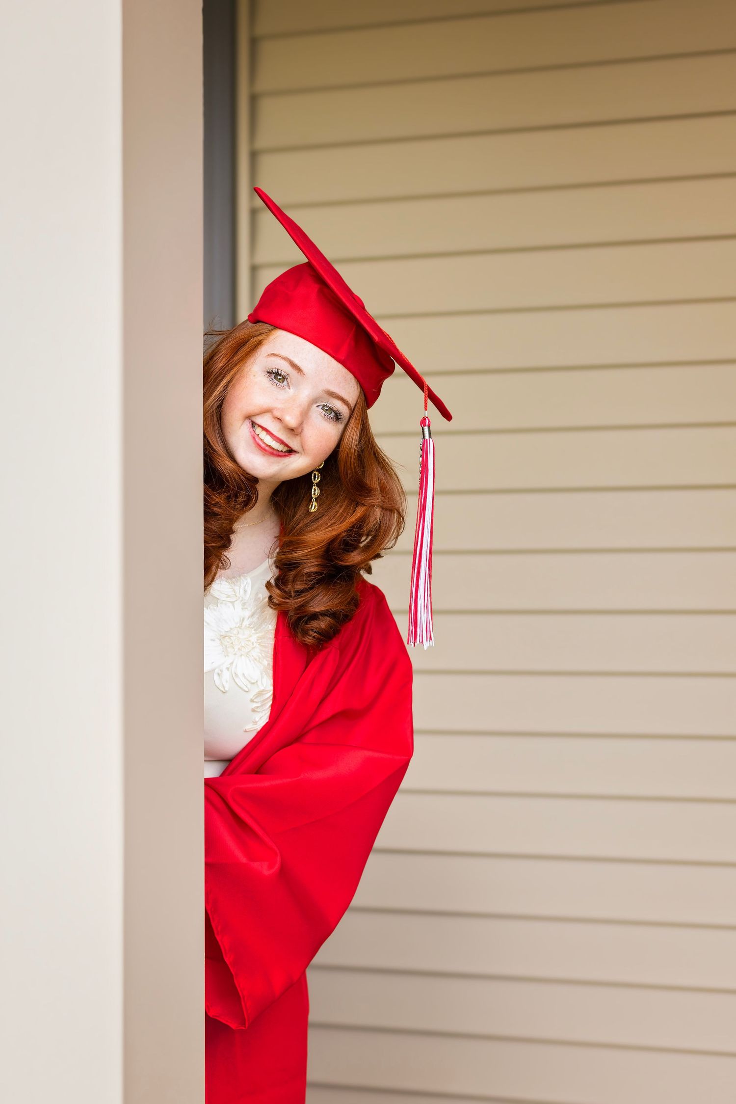 Happy redhead graduate peeking around corner wearing red cap and gown, smiling at camera.